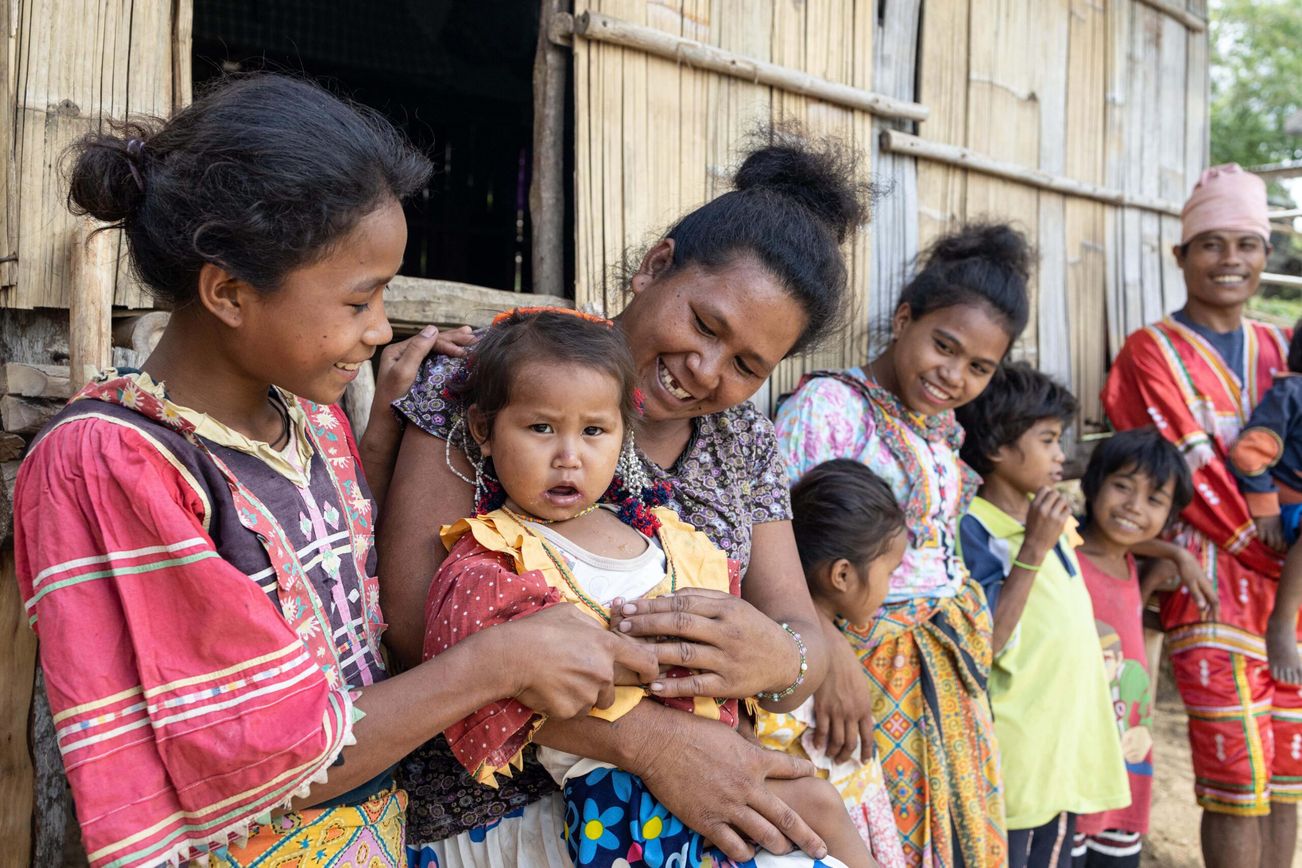 A group of young women stand in front of a wooden home while smiling at their children.