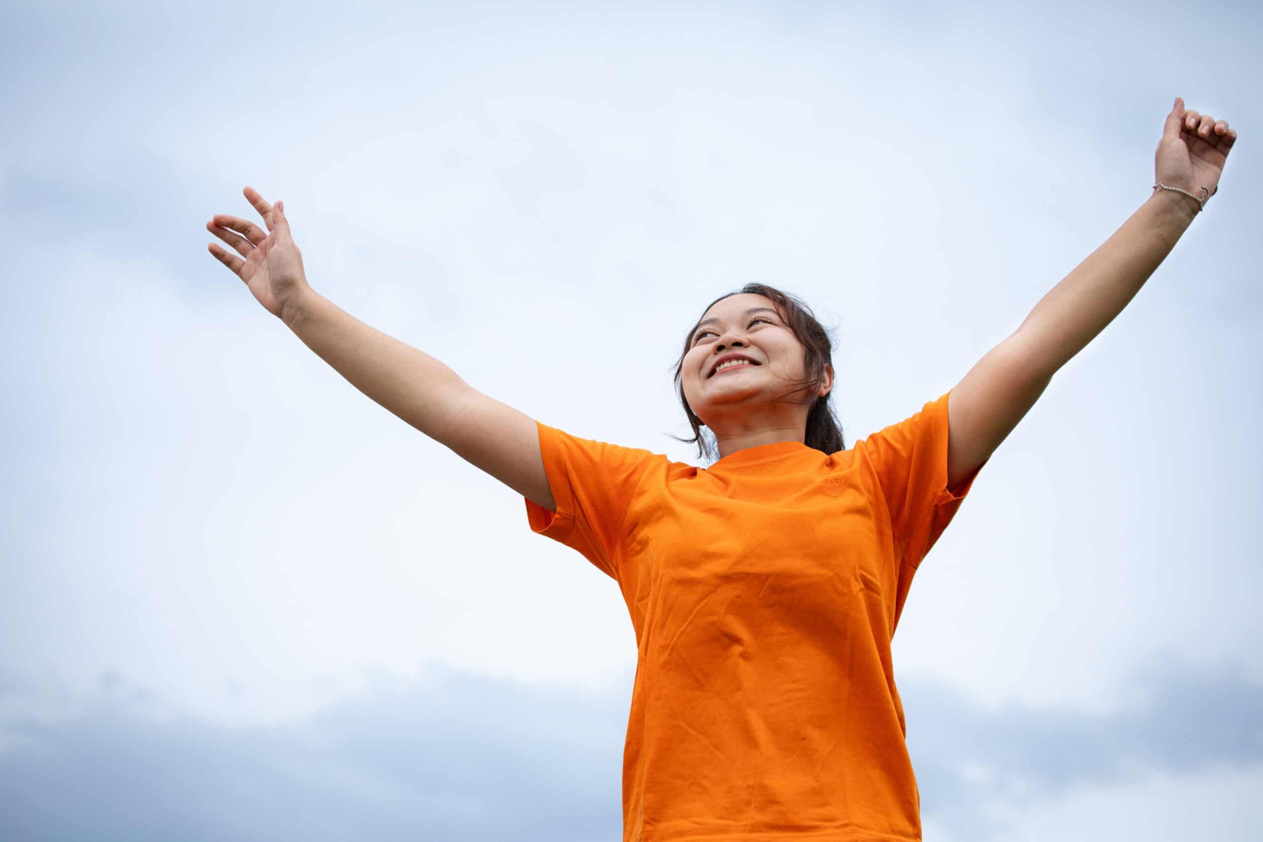 Young woman in a bright orange shirt stands with her hands raised while smiling.