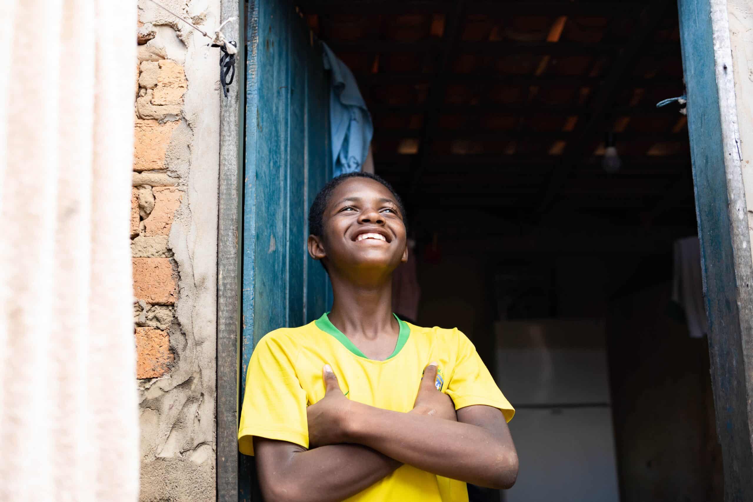 Young man wearing a yellow shirt leans on the door of his home while smiling and looking to the sky.