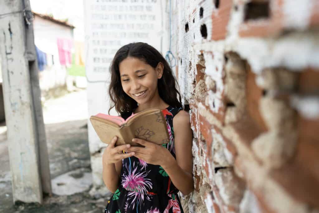 Young girl leans against a brick wall while reading her Bible and smiling.