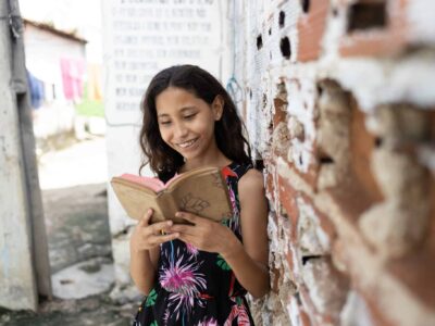 Young girl leans against a brick wall while reading her Bible and smiling.