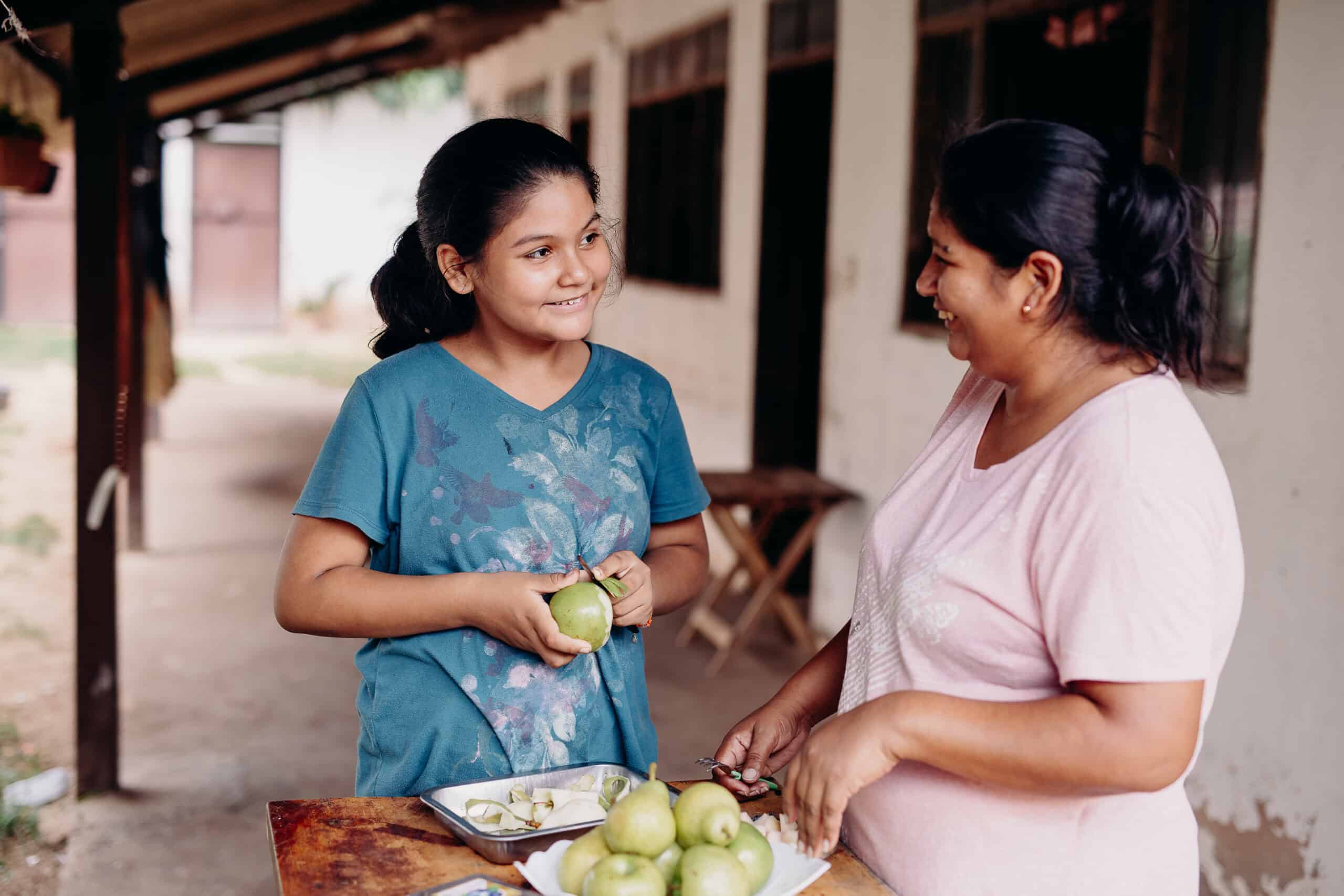 Young girl and her mother stand at a wooden table and peel apples while smiling at each other.