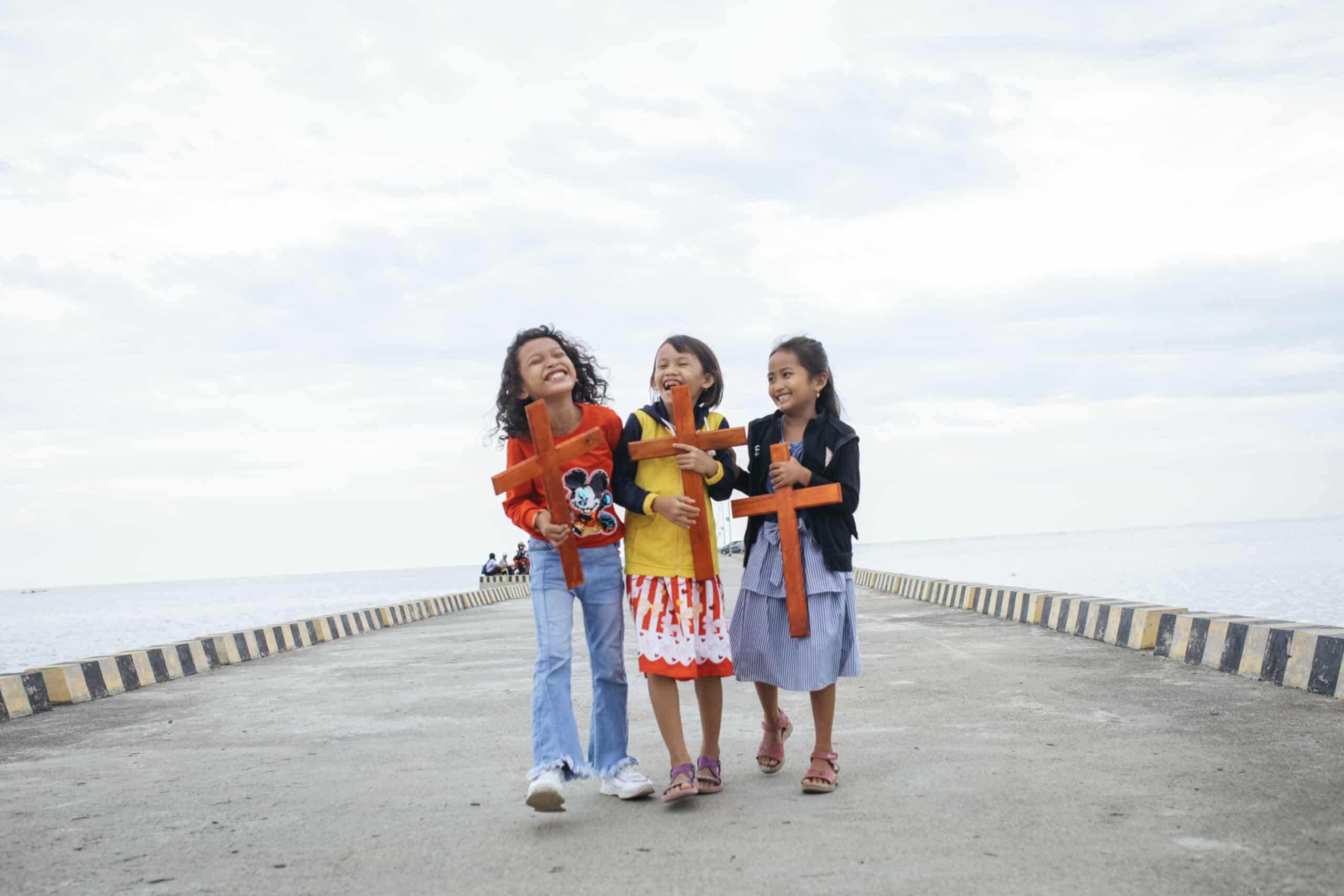 Three young girls stand on a dock holding wooden crosses and smiling.