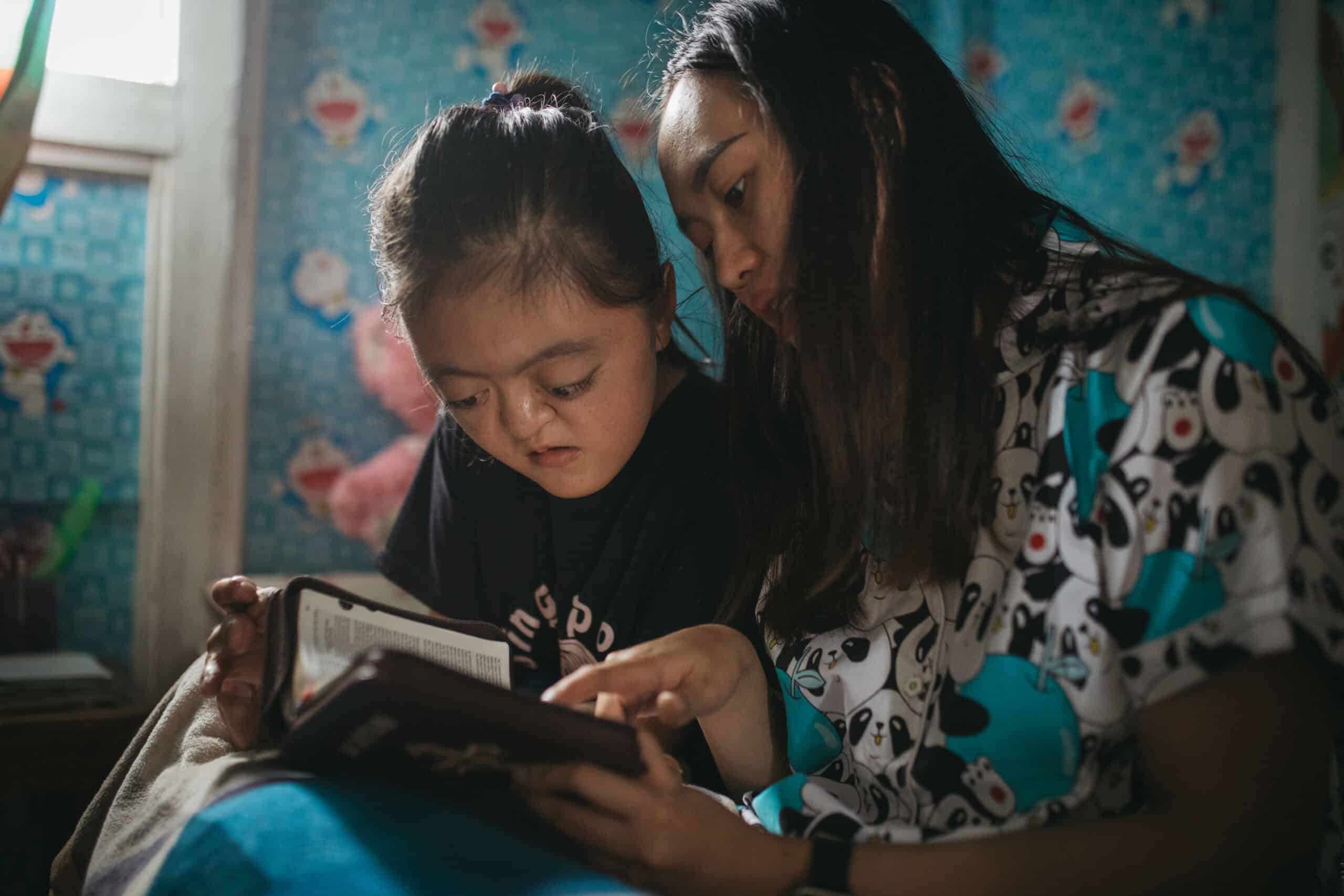 Young girl and her mother sit on her bed and read the Bible together.