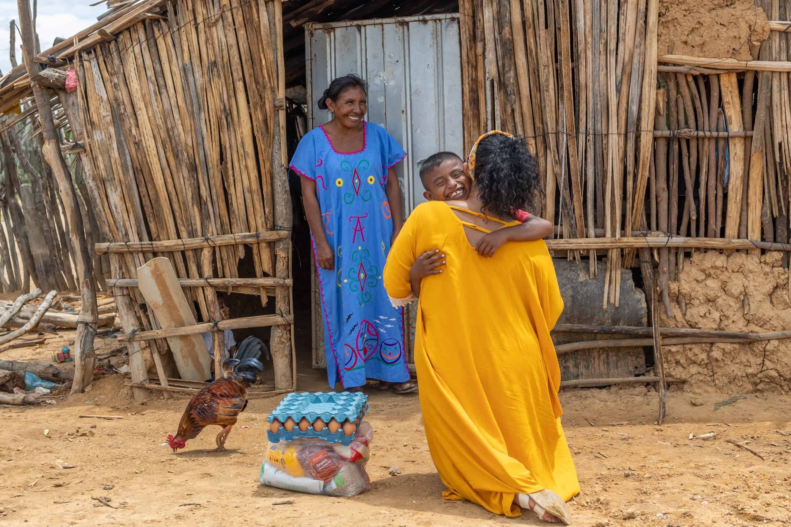 A young woman wearing a bright yellow dress hugs a young boy while kneeling next to a package of food.