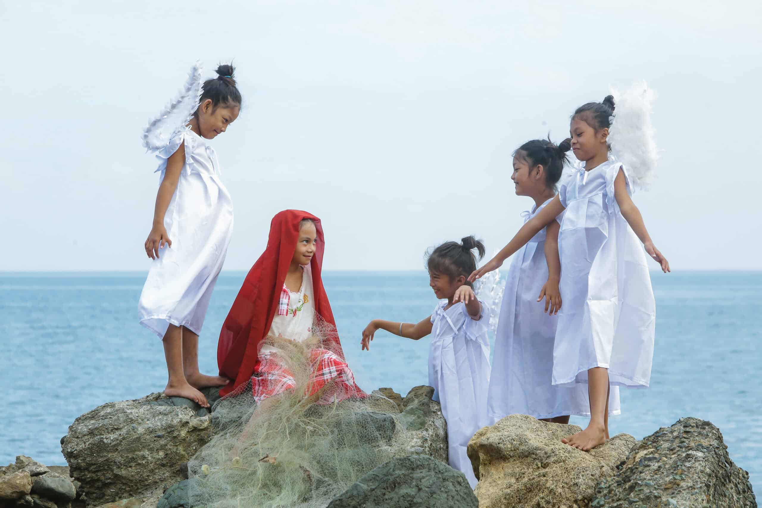 Four young girls wearing angel wings stand and smile at a young girl sitting on rocks and wearing a red headscarf.