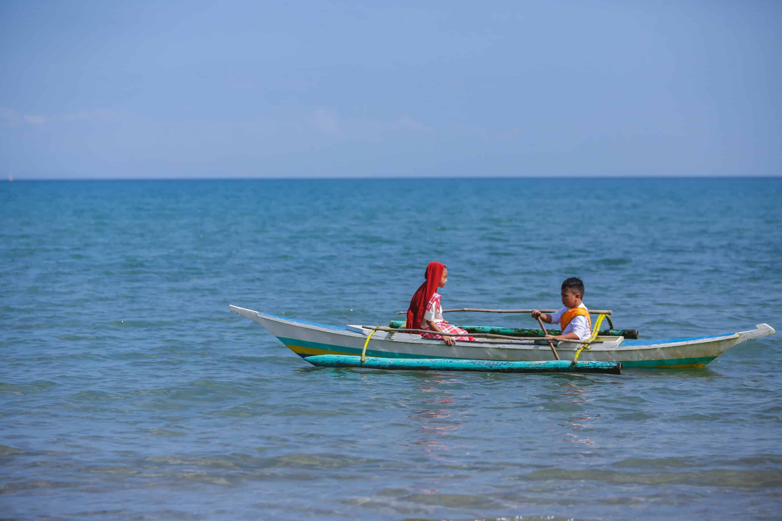 A young girl wearing a red headscarf and a young boy sit in a canoe and float on blue  water.