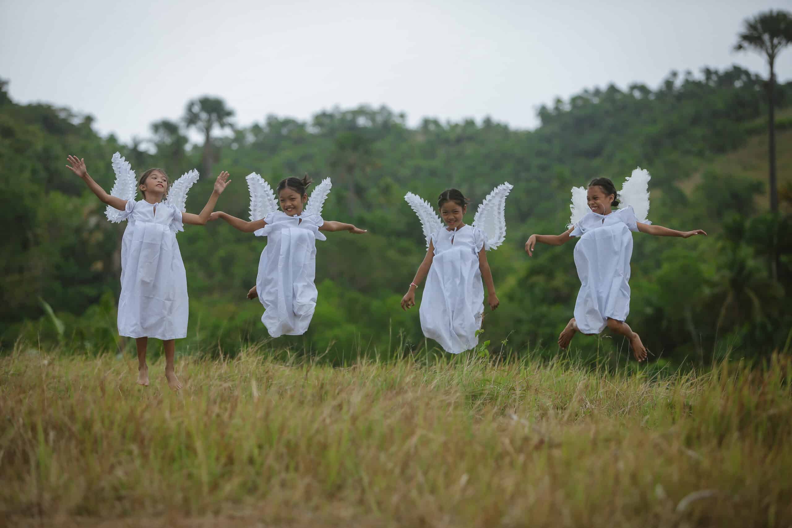 Four young girls jump in a field wearing white and angel wings.