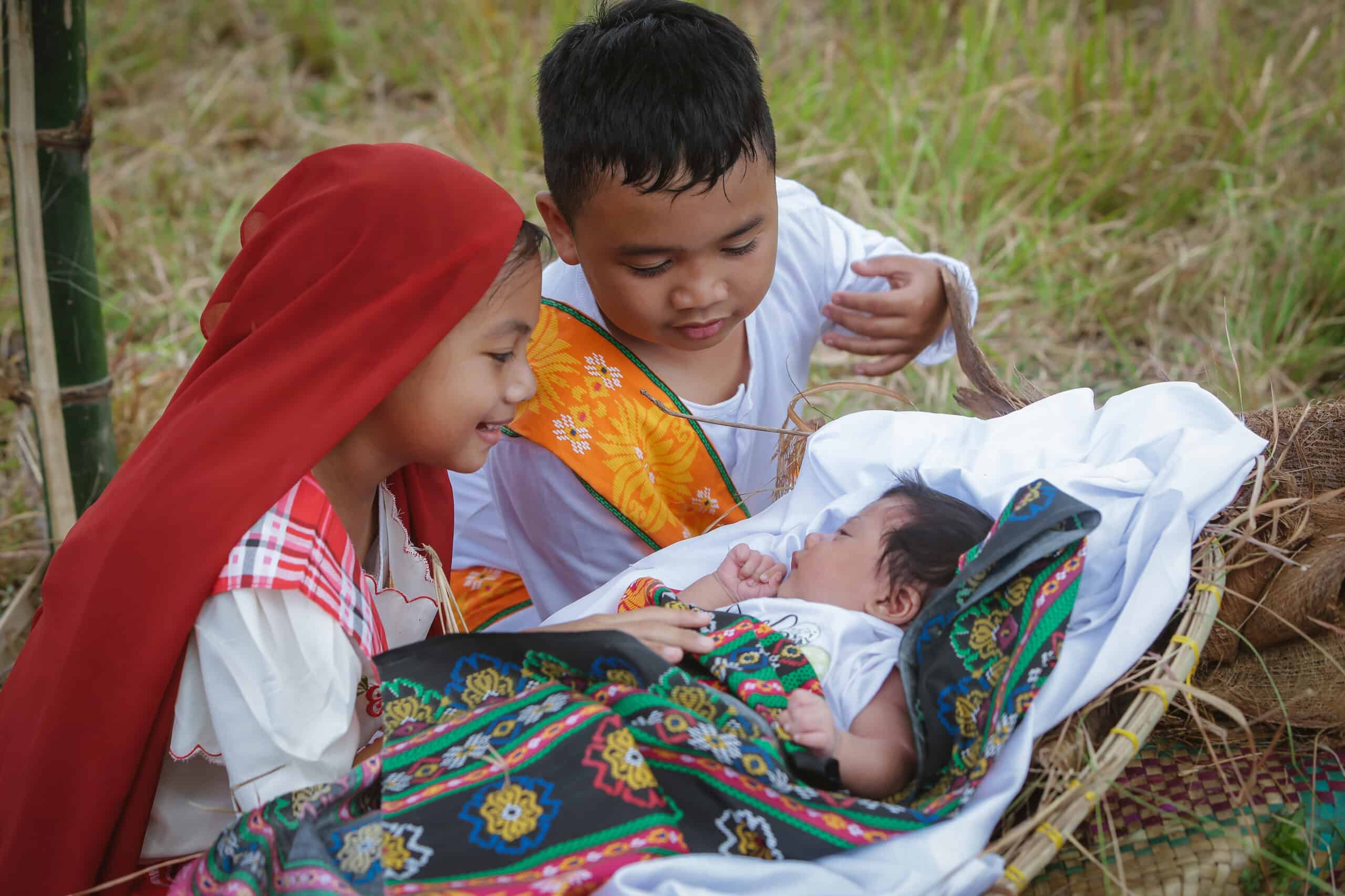 A young girl wearing a red headscarf sits next to a young boy. The girl holds a baby and smiles.