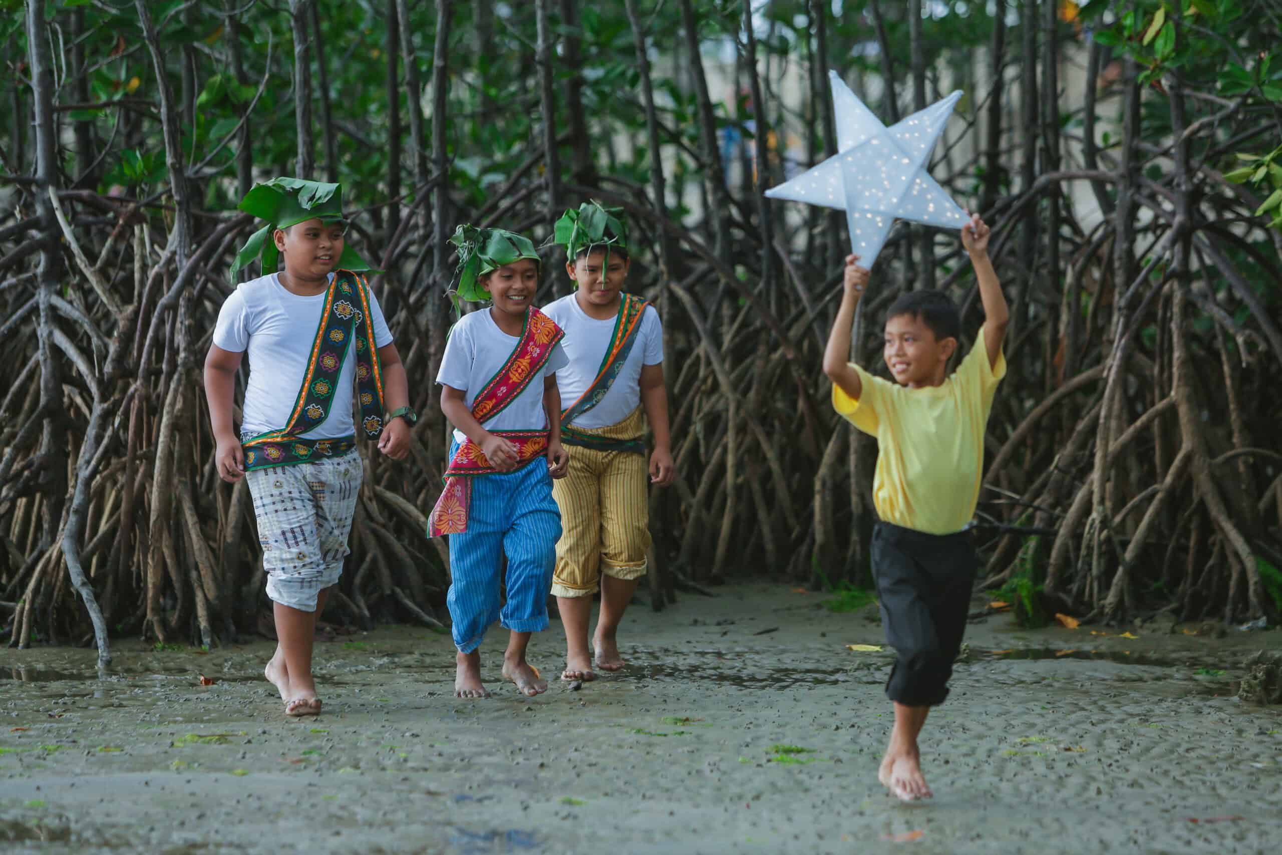 Four little boys run on the beach. They boy in the front holds a star above his head and smiles.