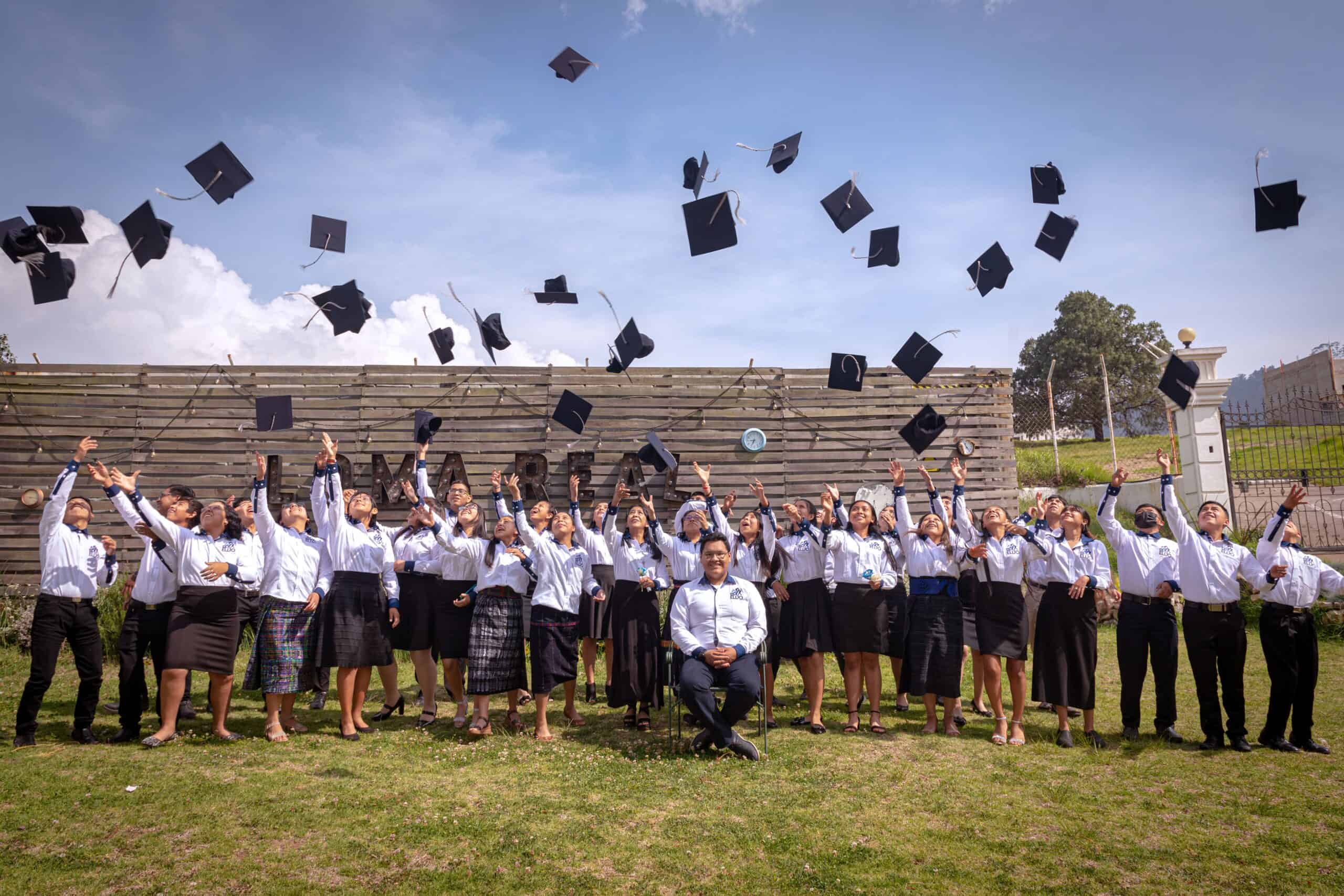 A group of young men and women stand in a field and throw graduation caps into the air.