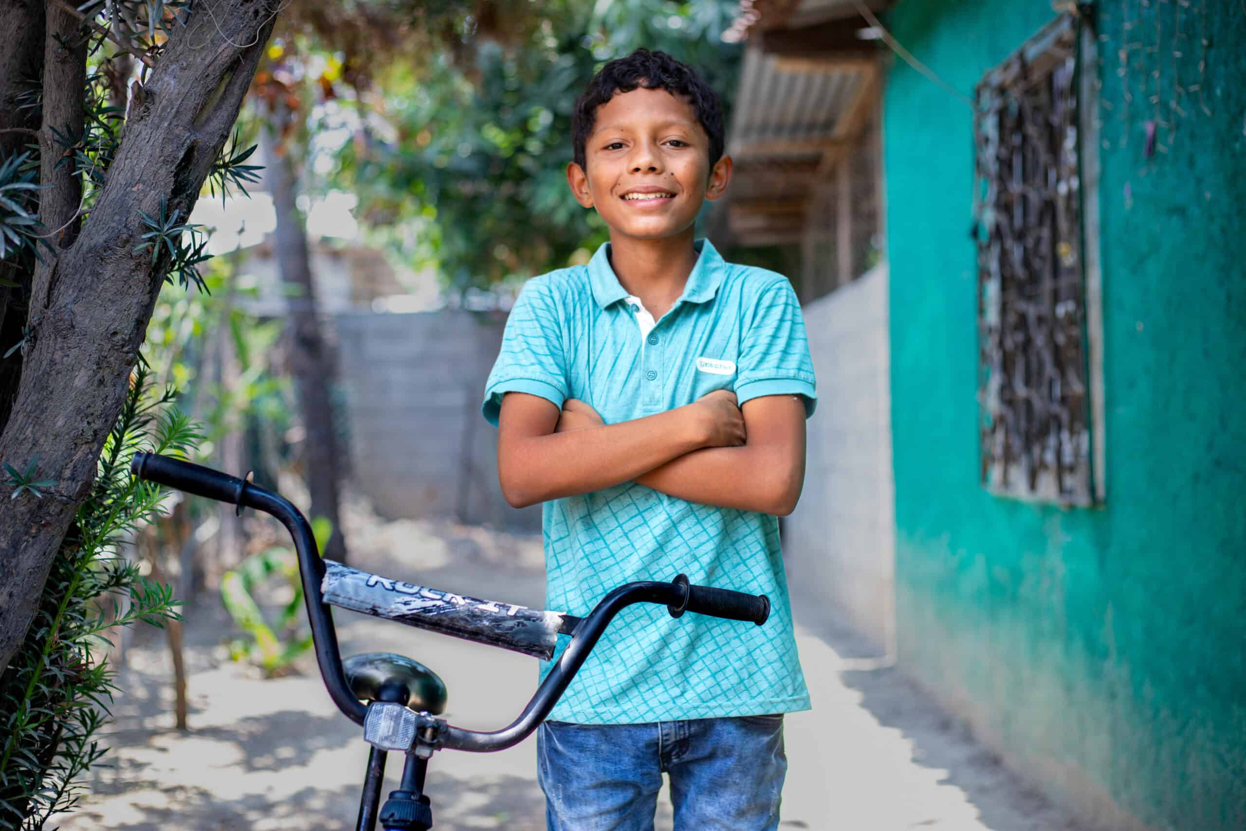 Denis Isai smiles at the camera while standing next to his bike, wearing a blur shirt and jeans, expressing gratitude for the support he received from the Compassion program in Honduras.