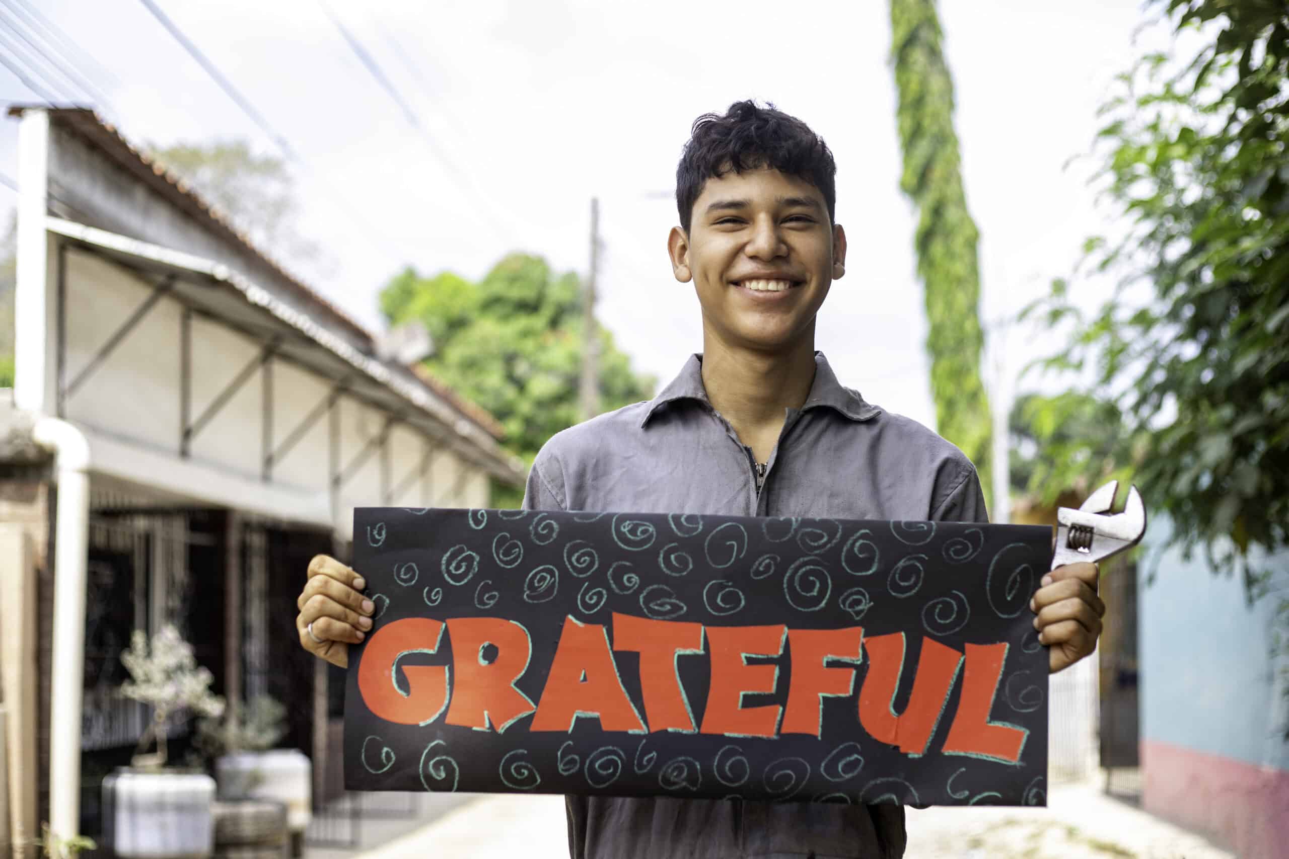 Brandon stands outside his house, smiling at the camera while holding a sign that says “Grateful” and a tool in his hand, showcasing his appreciation for support received from Compassion.