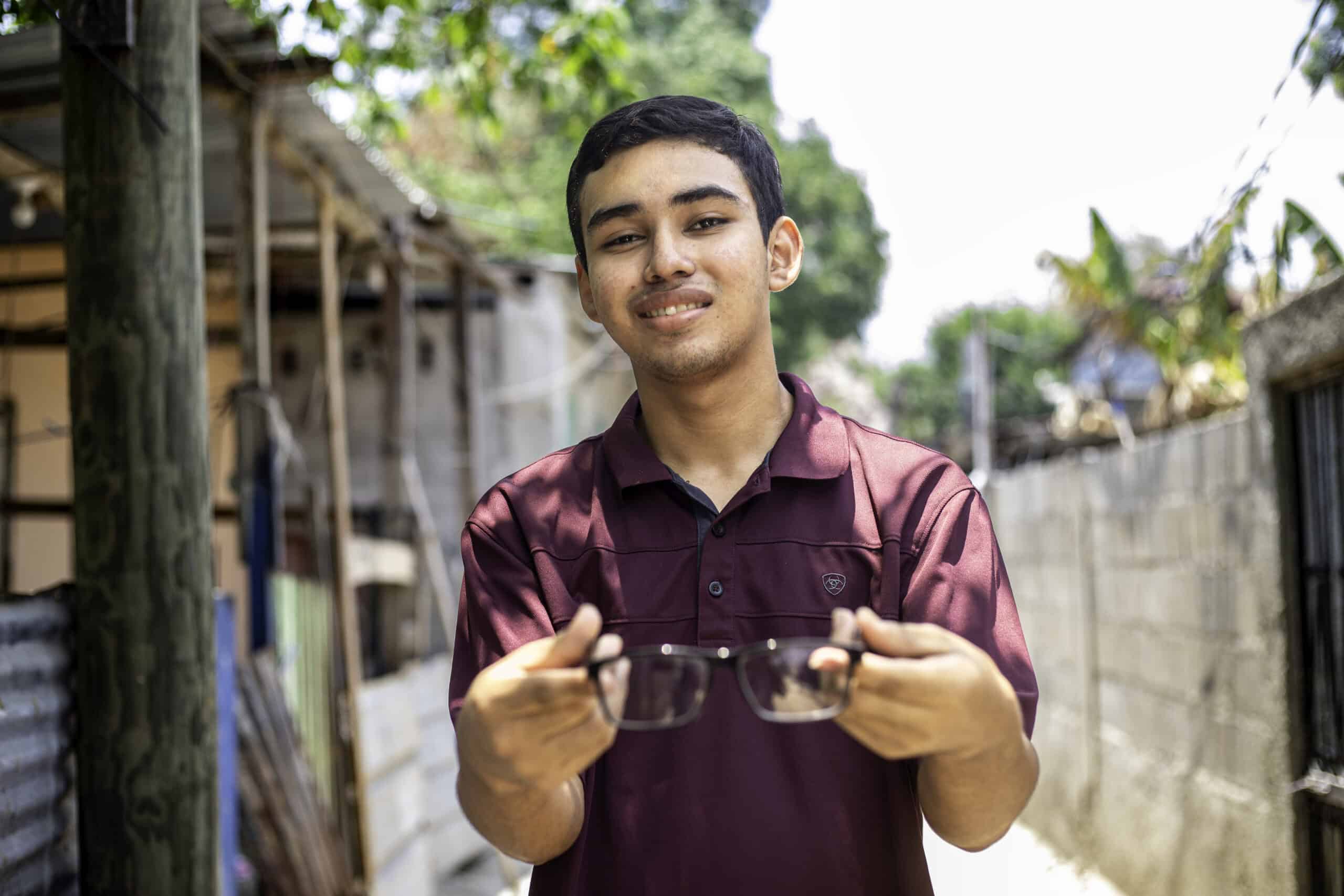 Angel smiles at the camera, holding the glasses he received from the Compassion center while wearing a red and purple shirt.