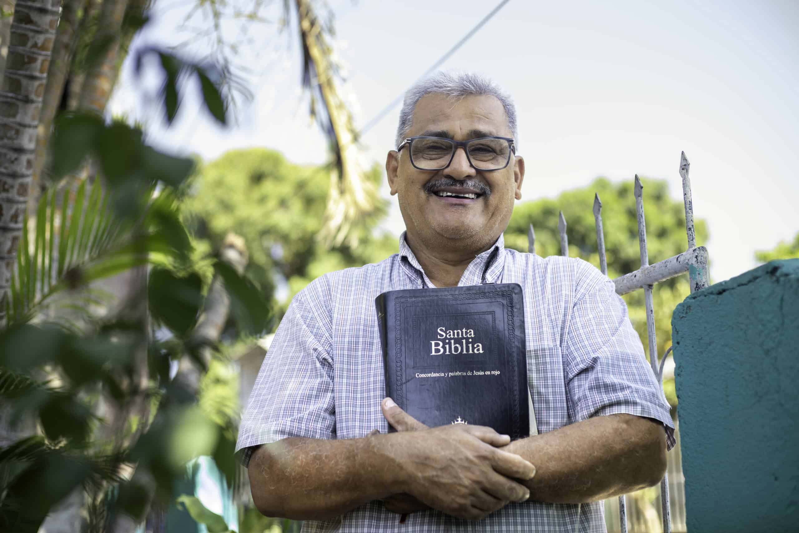 Santos, the Center Director, stands outside his house, smiling at the camera while holding a Bible.