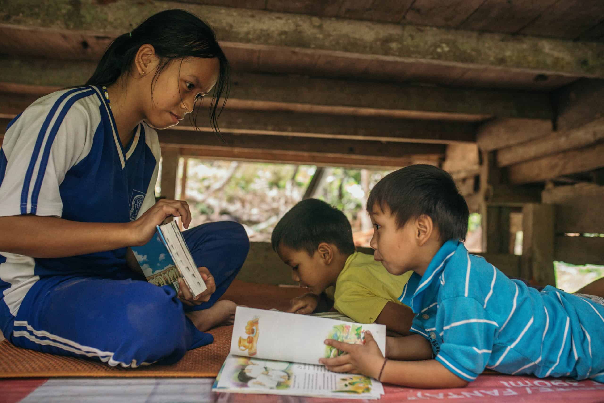 A young boy wearing a striped shirt lies on the floor while flipping through a Bible. A young woman sits in front of him holding another Bible.