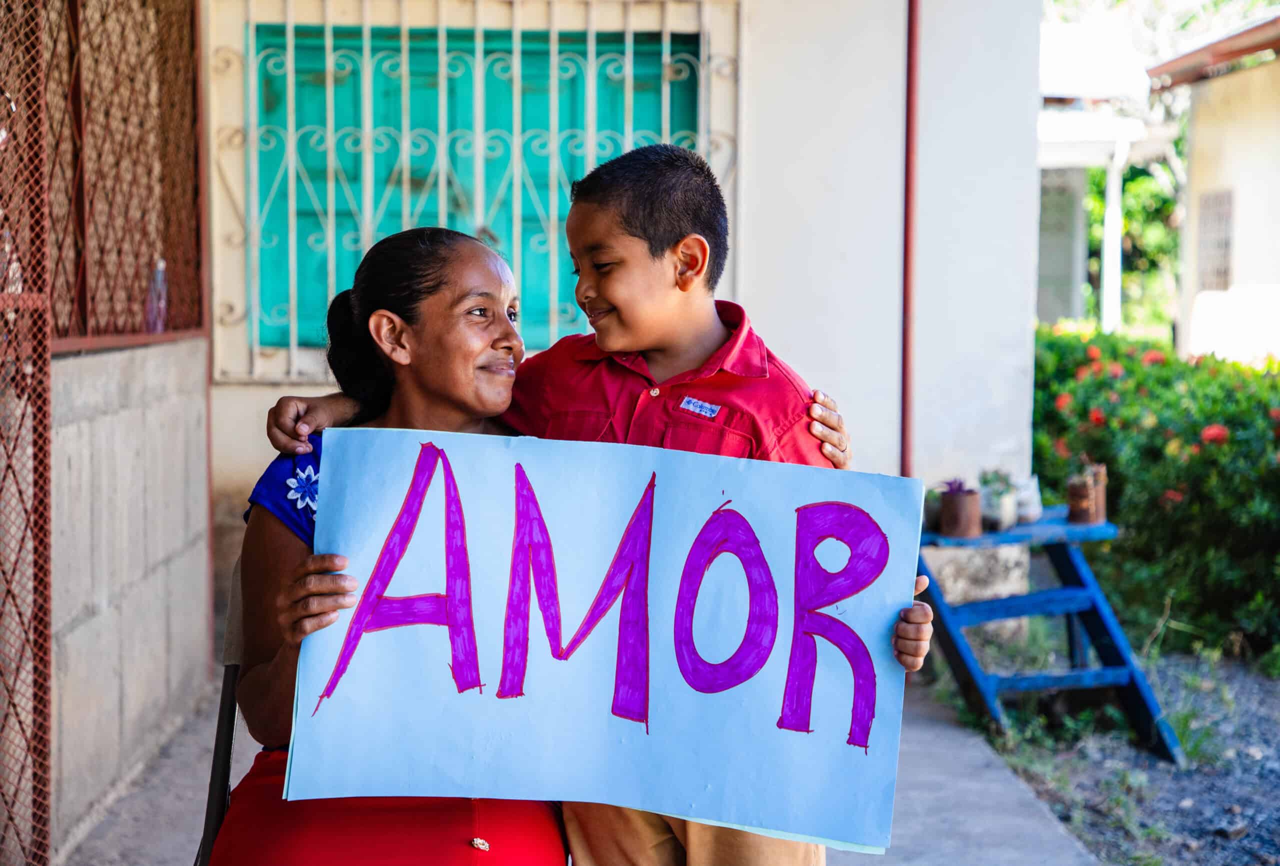 Woman and child have their arms around each other holding a sign that says "amor". 