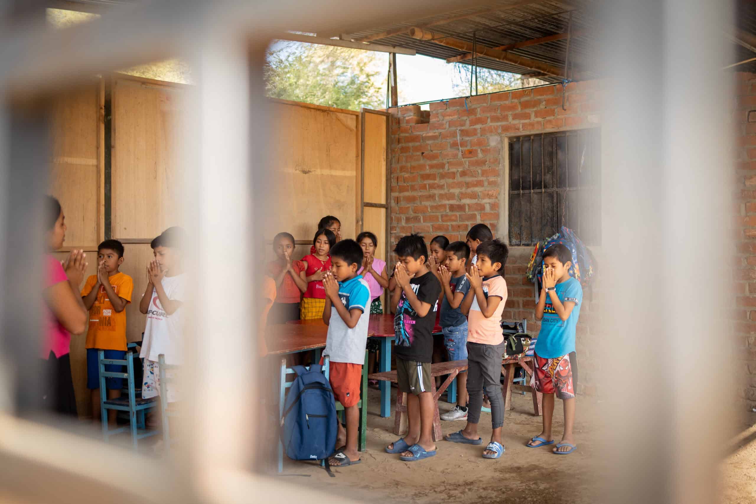 A group of children stand with their hands clasped in front of their faces inside of a brick classroom.