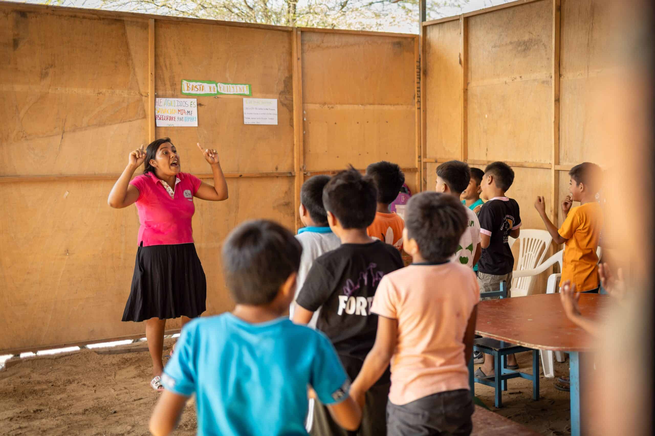 Young woman wearing a bright pink shirt stands in front of a classroom of children with her hands raised in the air as she sings.