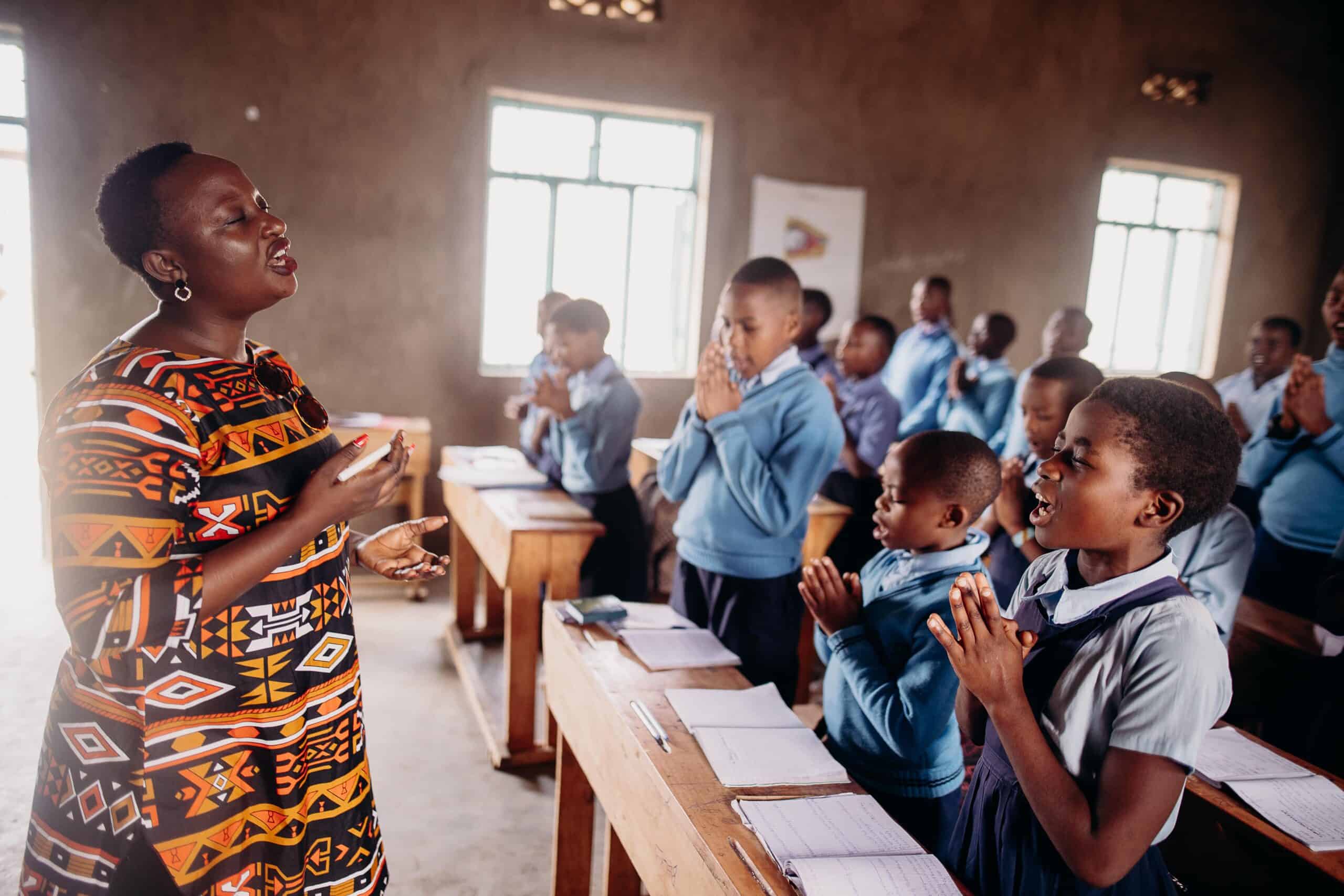 A woman stands in front of a classroom wearing a patterned dress. She has her eyes closed and is leading the children through a prayer. The children stand with their eyes closed and hands clasped.