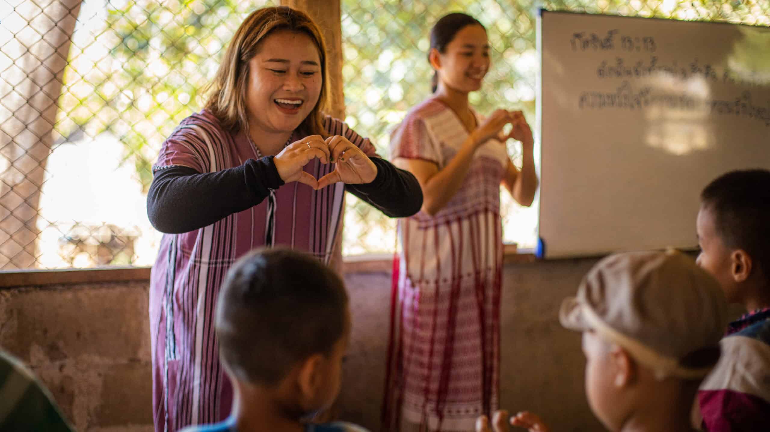 A teacher stands in front of the classroom smiling and holding her hands in the shape of her heart. 