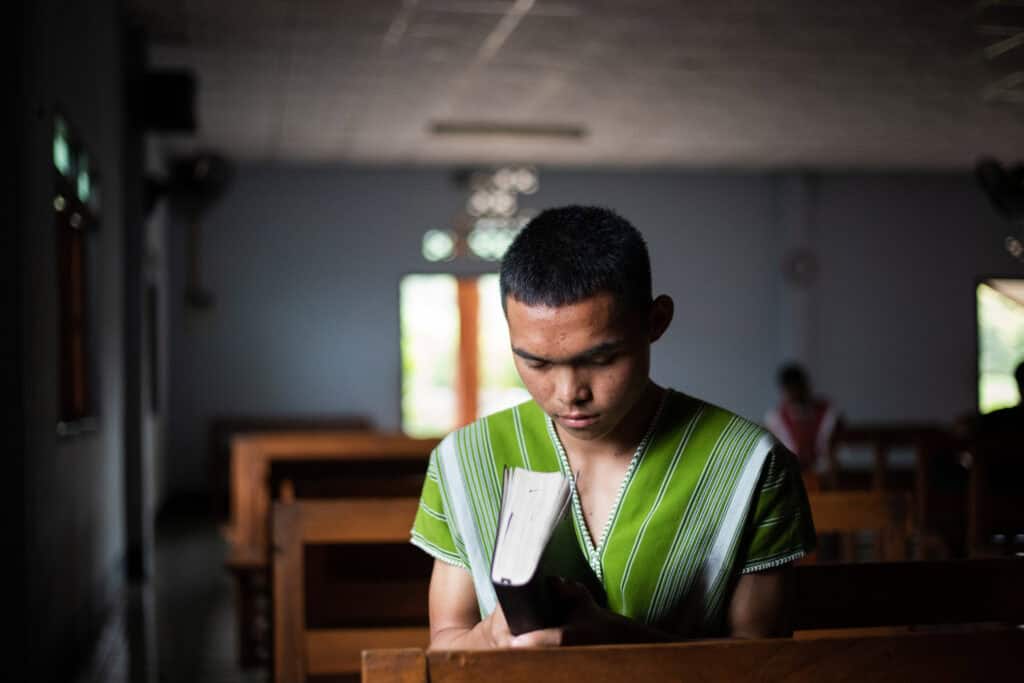 A young man sits amid church pews holding a Bible with his head bowed and eyes closed. How to grow closer to God: 10 Ways