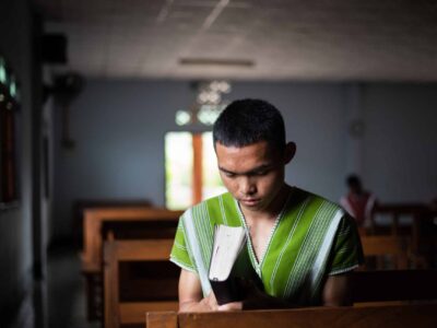 A young man sits amid church pews holding a Bible with his head bowed and eyes closed. How to grow closer to God: 10 Ways