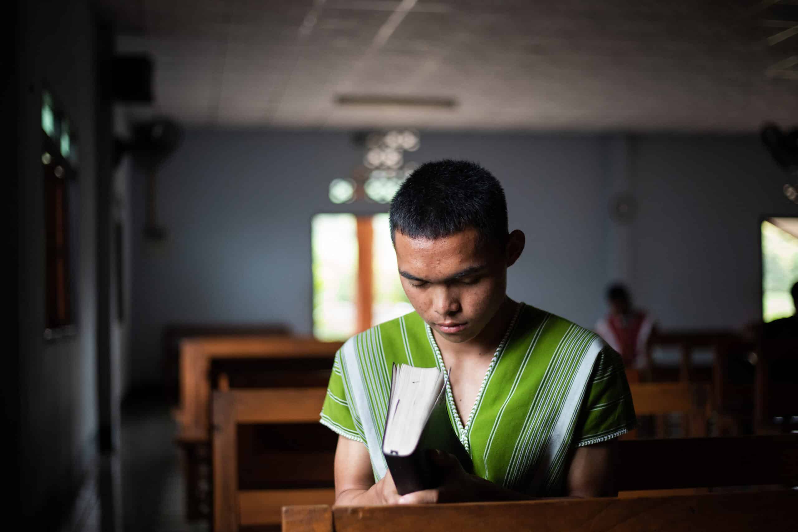 A young man sits amid church pews holding a Bible with his head bowed and eyes closed. How to grow closer to God: 10 ways.