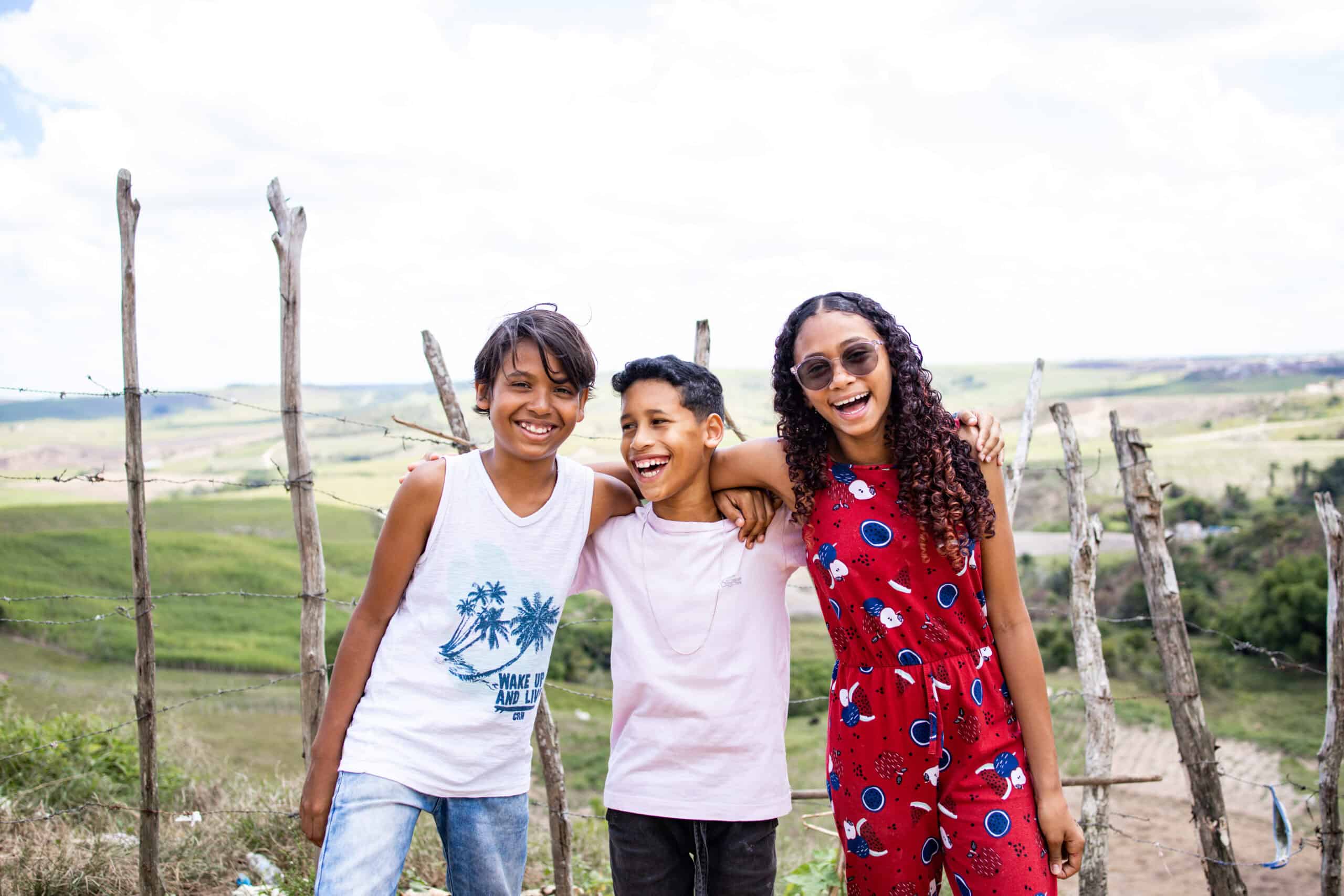 Three kids stand arms around each other facing the camera and smiling.