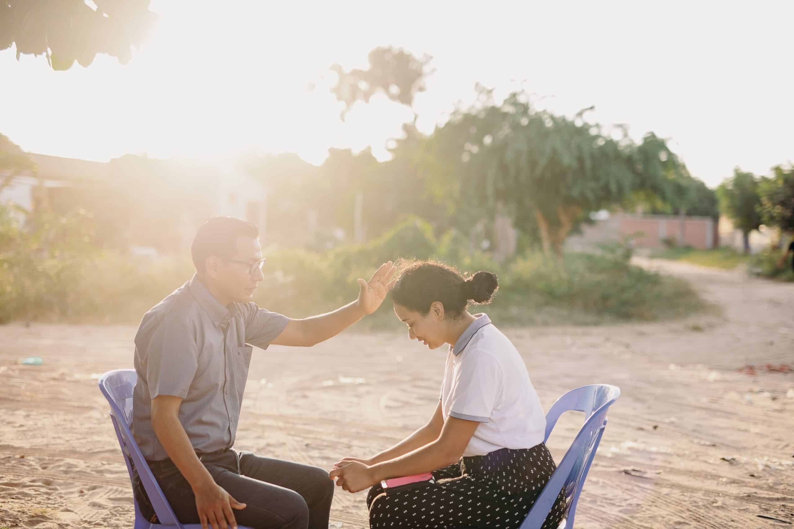 A young woman sits in front of a man outside with her eyes closed and head bowed. The man holds his hand in front of the woman’s head with his eyes closed. They pray with sunlight coming in from the upper lefthand corner.