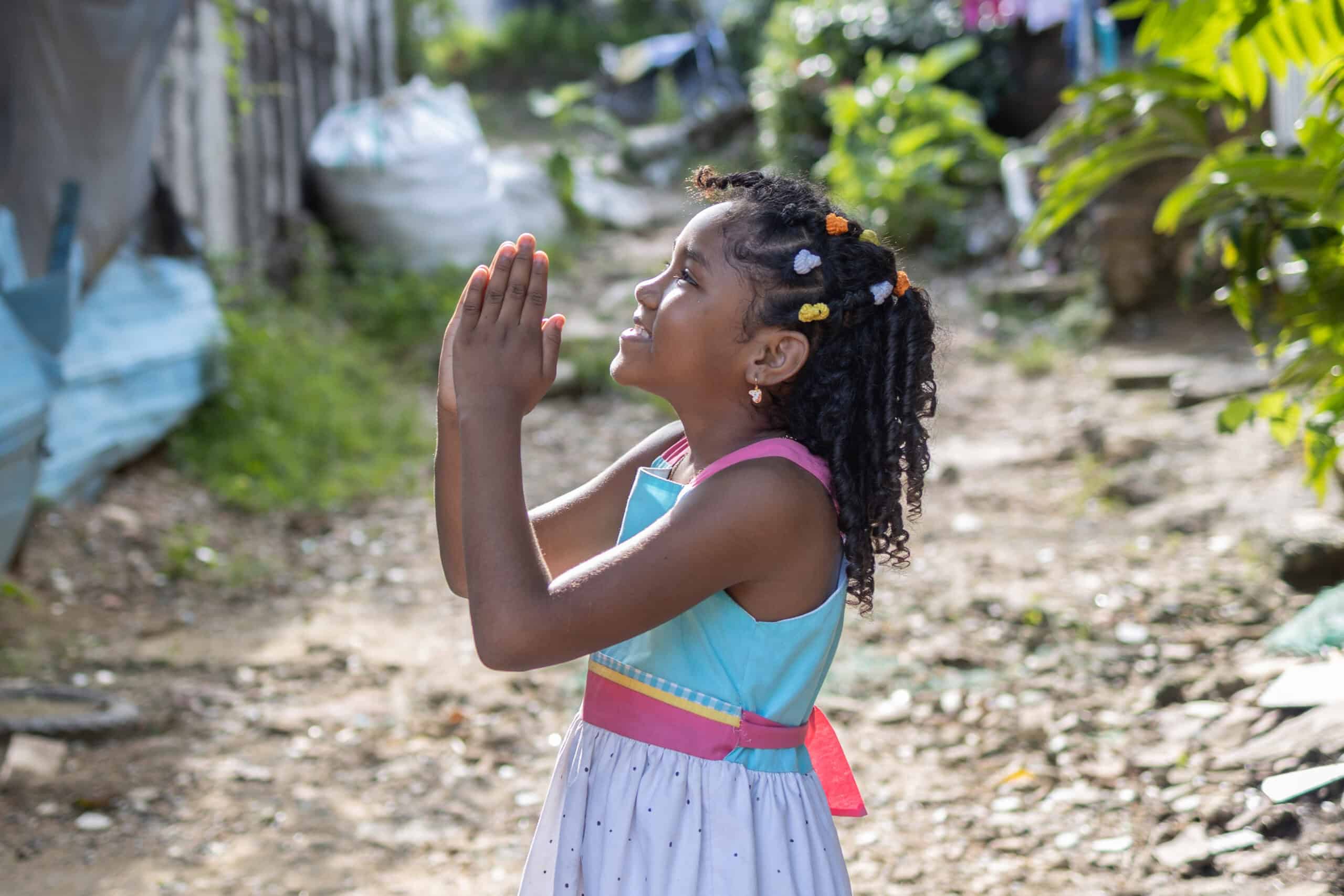 Young girl wearing a colorful dress stands with her head raised to the sky with her hands clasped together as she smiles.