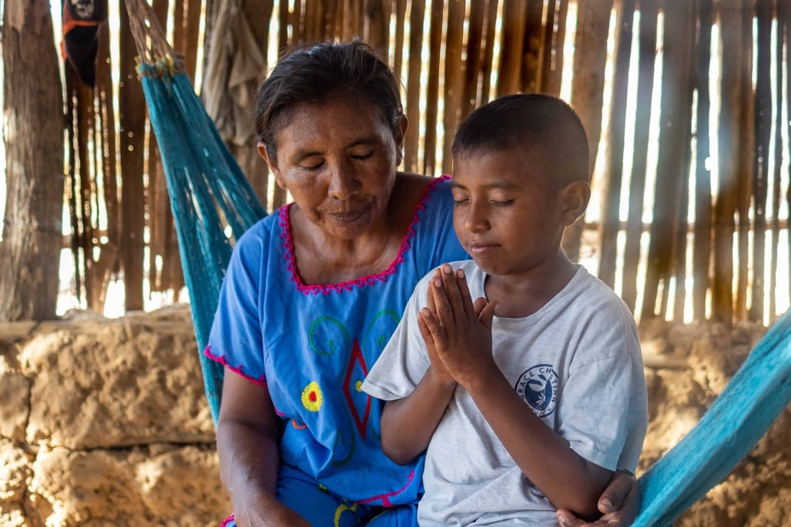 A young boy and a woman bow their heads in prayer. The boy holds his hands in front of his heart.