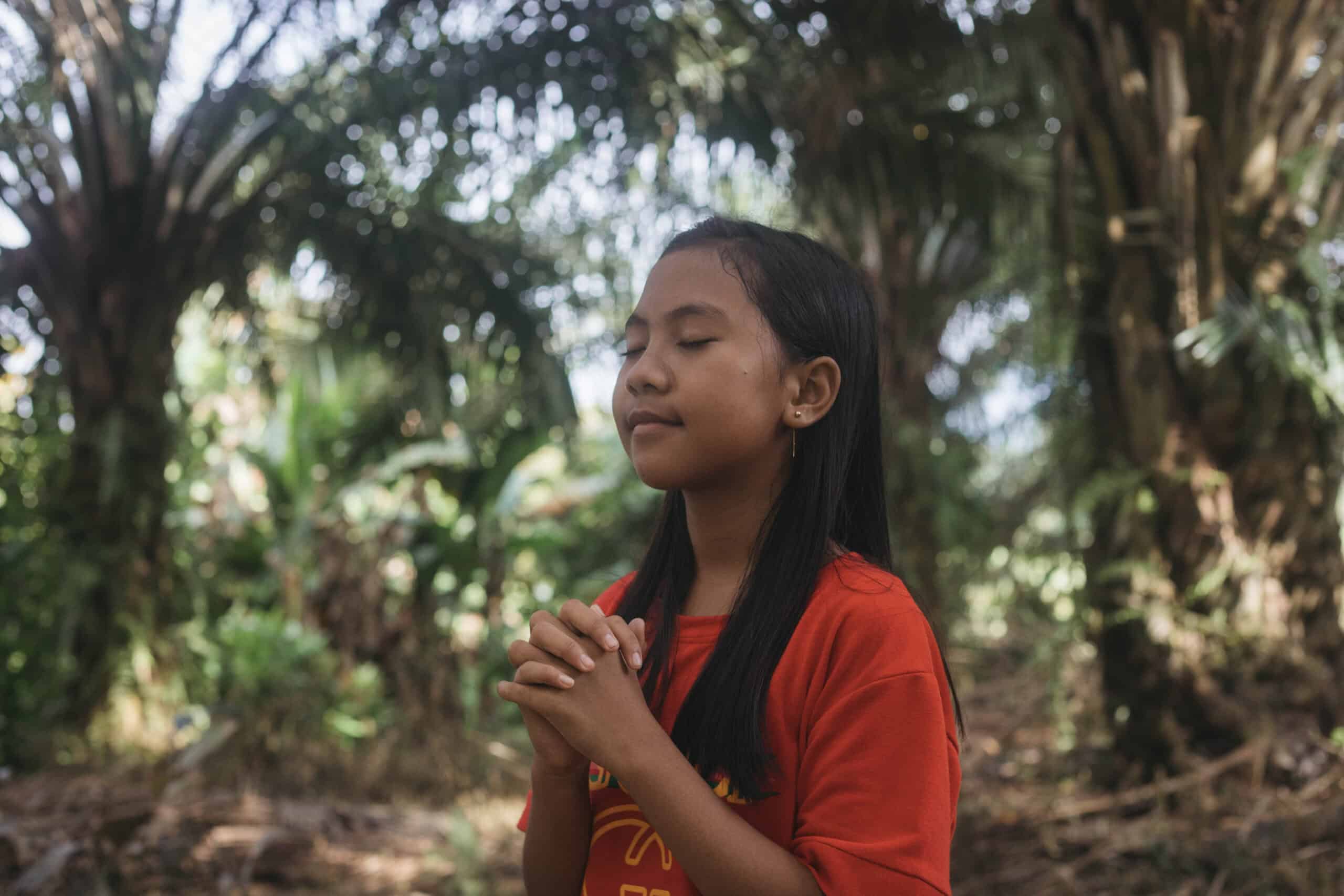 A young girl folds her hands at her chest and close her eyes to pray while surrounded by trees.