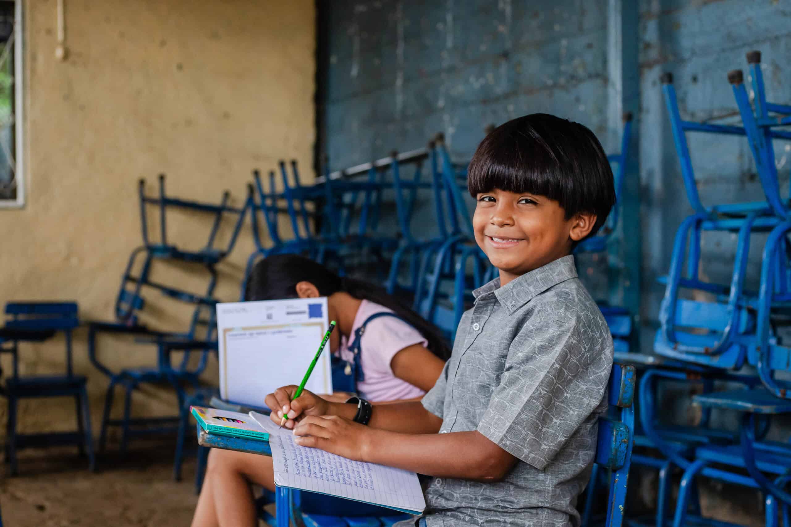 Charlie is writing the first draft of his sponsor letter in his notebook at a desk surrounded by colored pencils. He looks up at the camera with a smile.