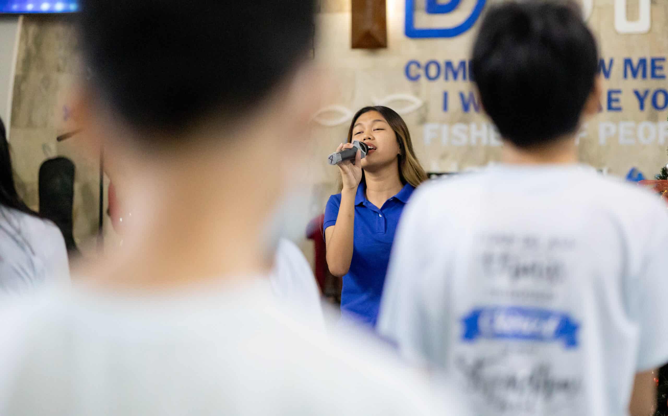 A teenage girl wearing a bright blue shirt sings into a microphone.