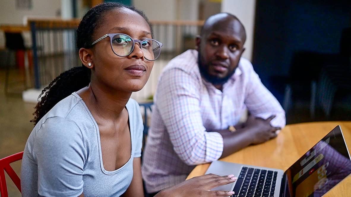 Sekata and Pacifique, the project director, are seated in front of a laptop to watch a video of Sakata’s mobile application, BALDI.]