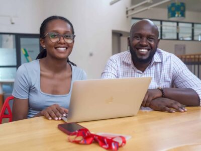 A young woman sits next to an older man. They both smile for the camera in front of a computer.