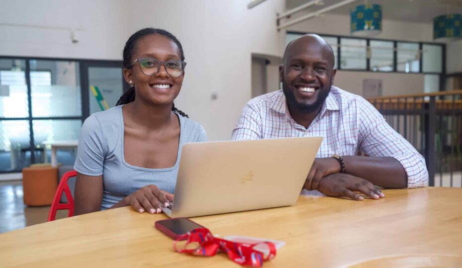 A young woman sits next to an older man. They both smile for the camera in front of a computer.