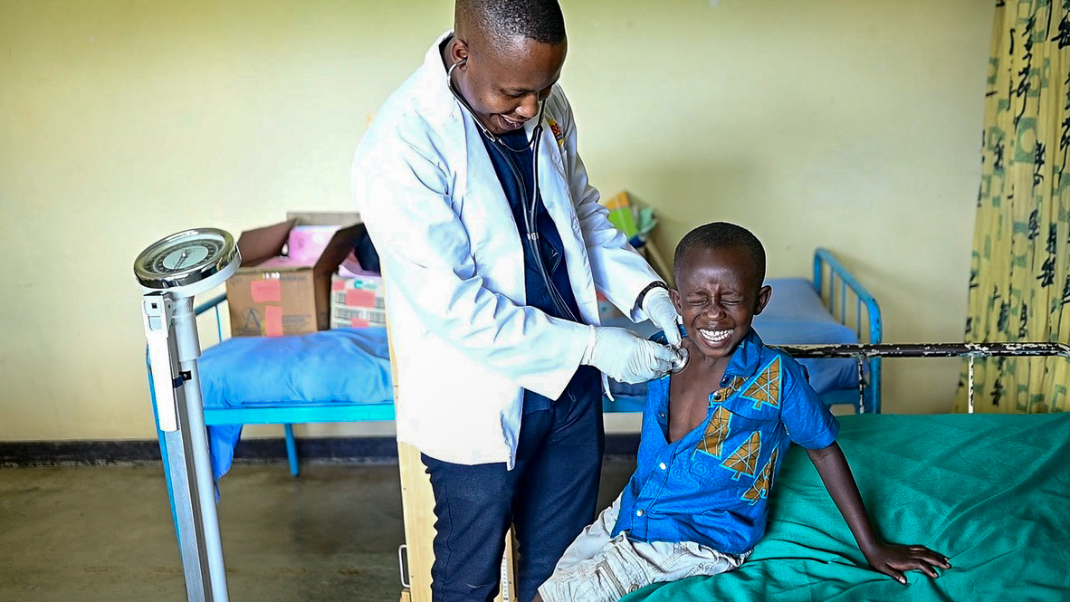 A young boy sits on a hospital bed while a doctor checks his heart. The boy smiles brightly for the camera.