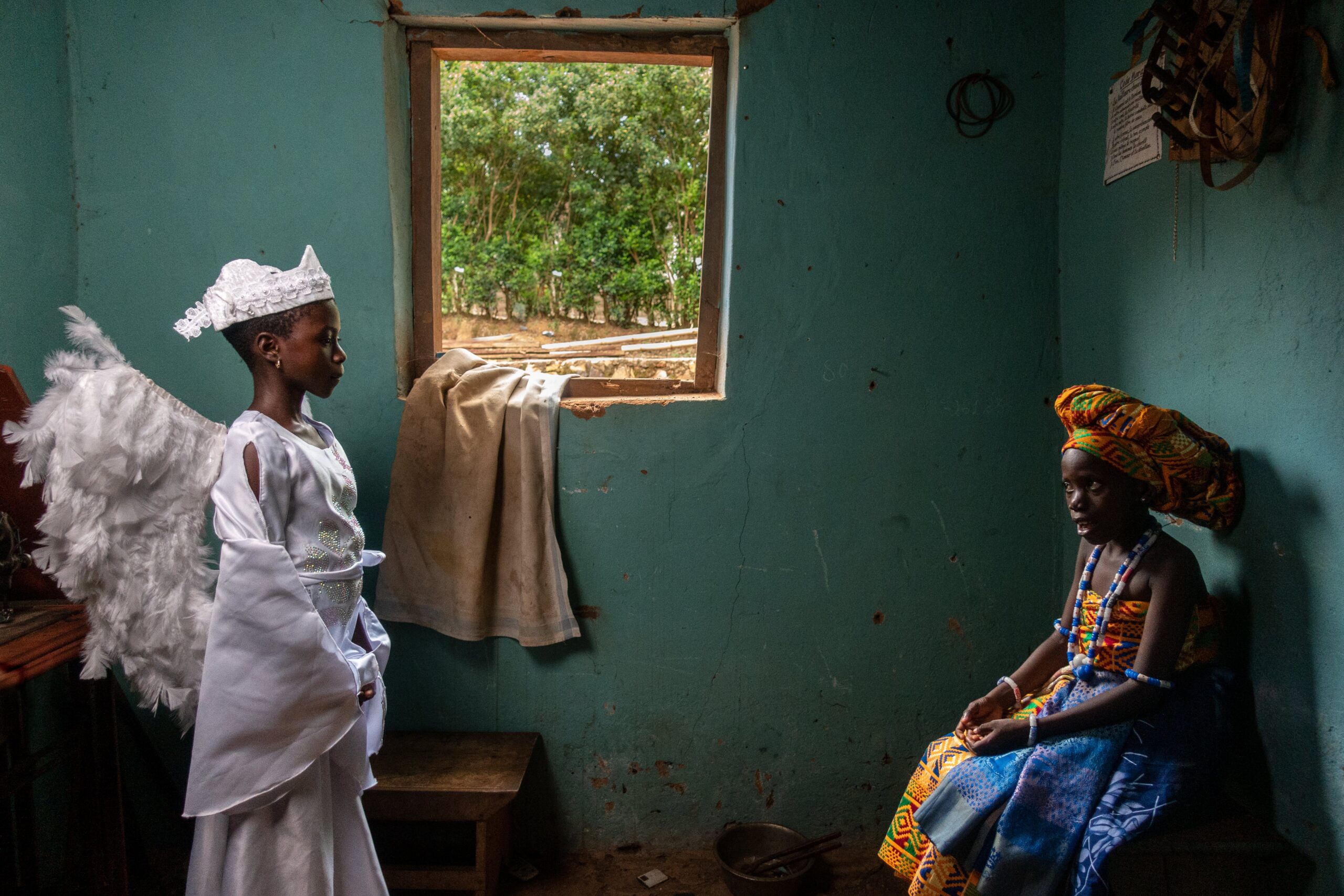 CC-TG0946-Nativity-010-2506-min A girl dressed as an angel stands across the room from a girl dressed in traditional dress, with a window between them.