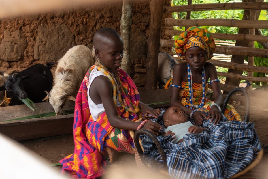 A boy and girl kneel around a basinet and a baby in a stall with goats in the background.