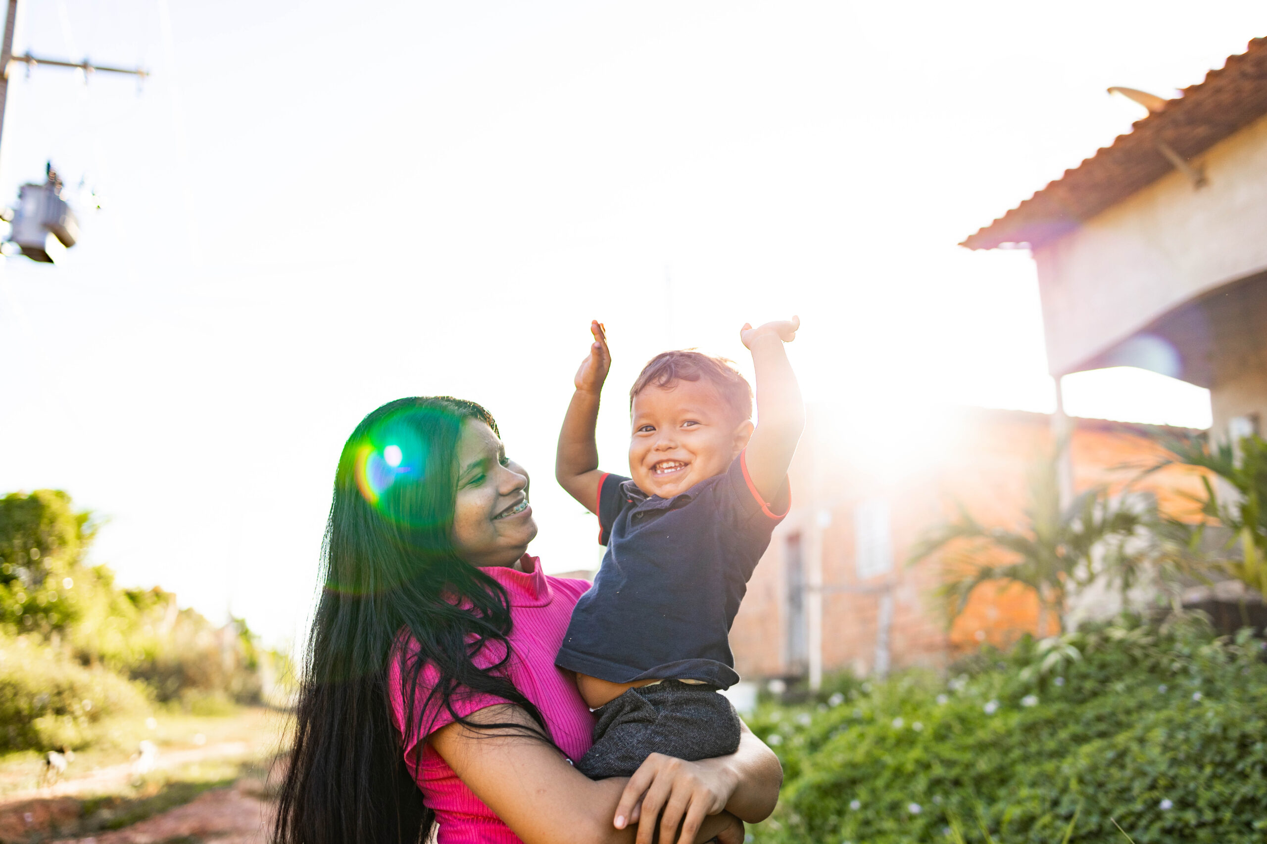 Woman holds a young boy who has his arms up in the air and is smiling bright for the camera.