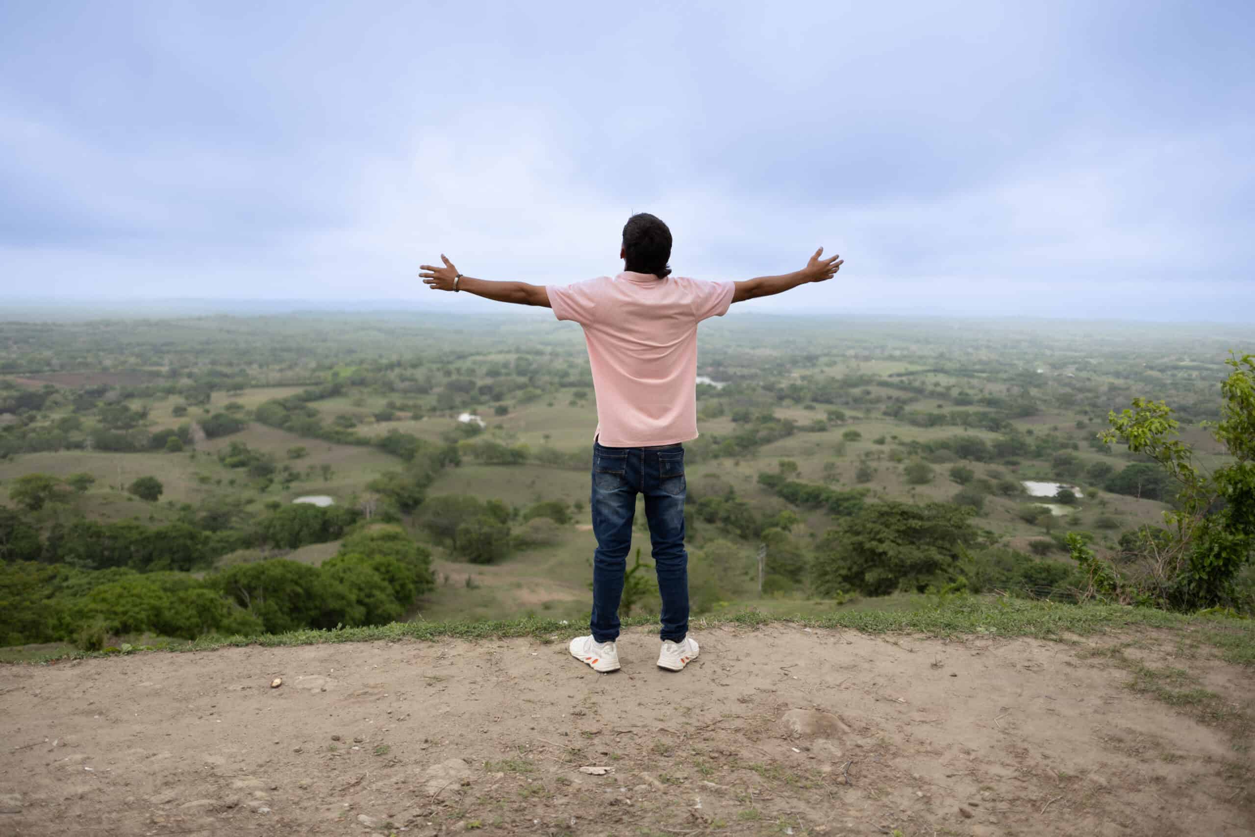 A young man wearing a pink shirt stands on a hill with his back turned and arms lifted to the sky.