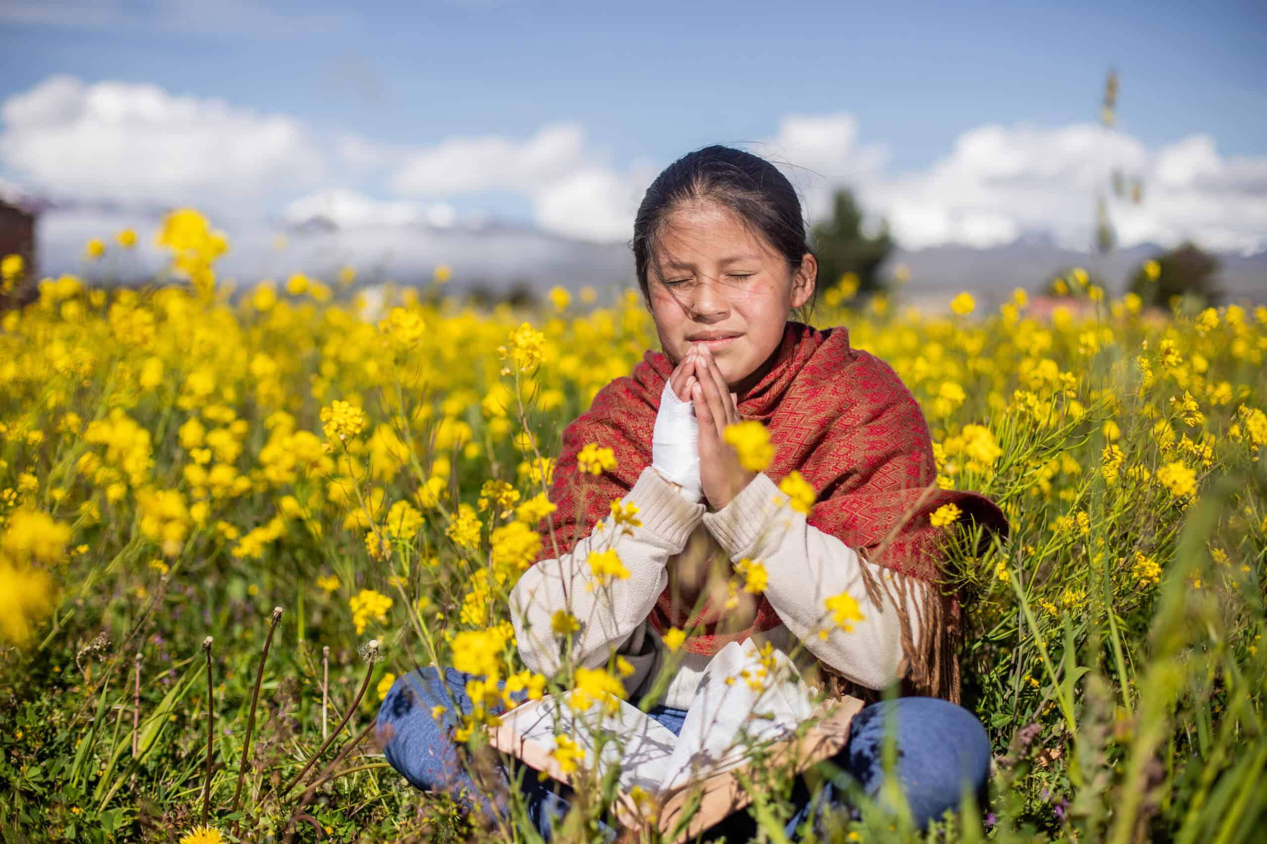 Young girl sits in a field of yellow flowers with her eyes closed and hands together as she prays.