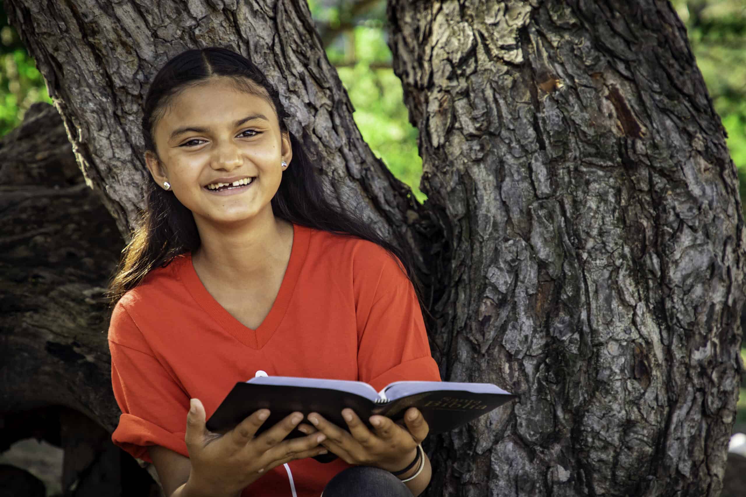 A girl in a red shirt sits in a tree holding a Bible and smiles at the camera.