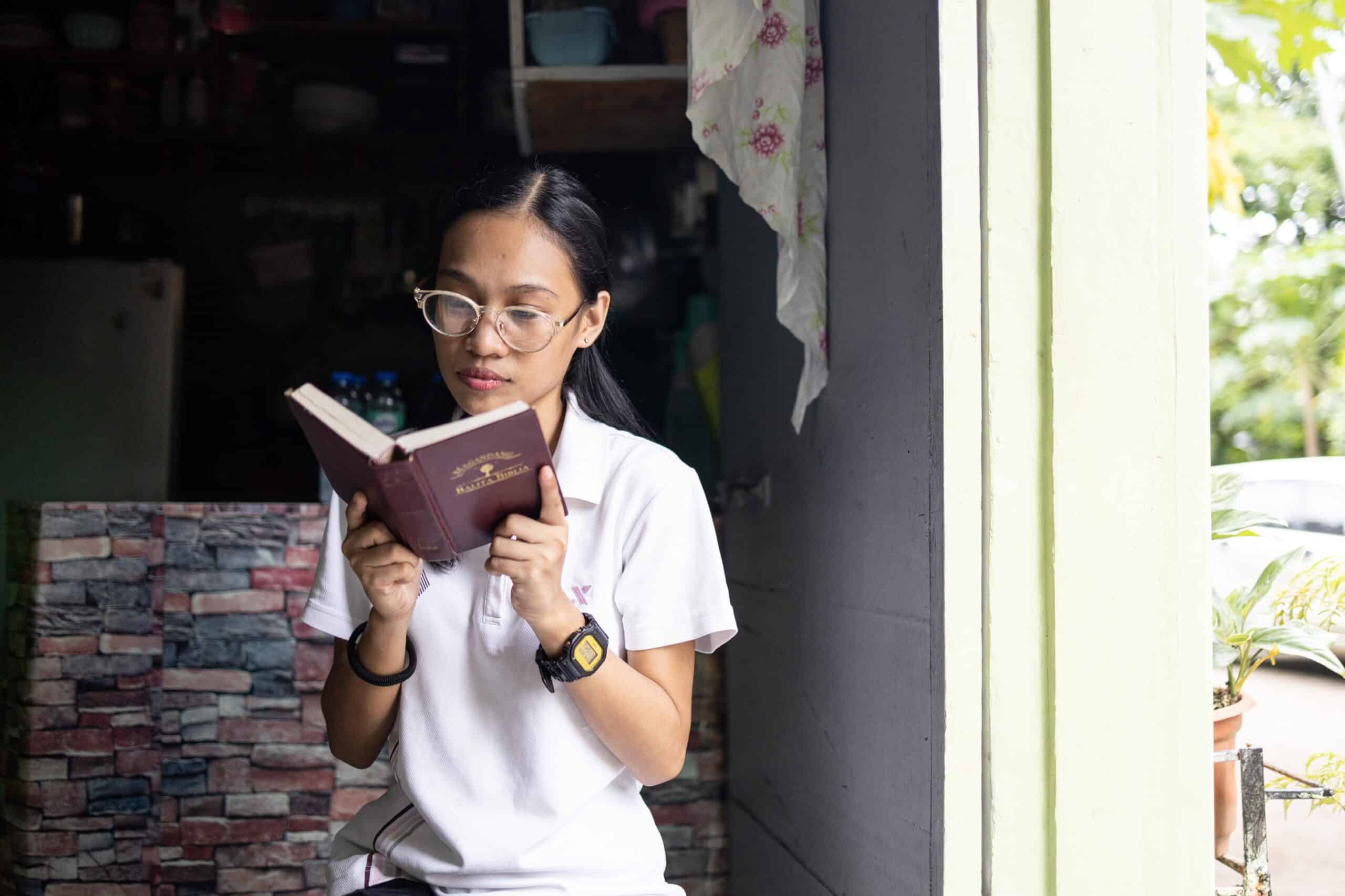 Woman wearing a white polo stands near a doorway while reading a Bible.