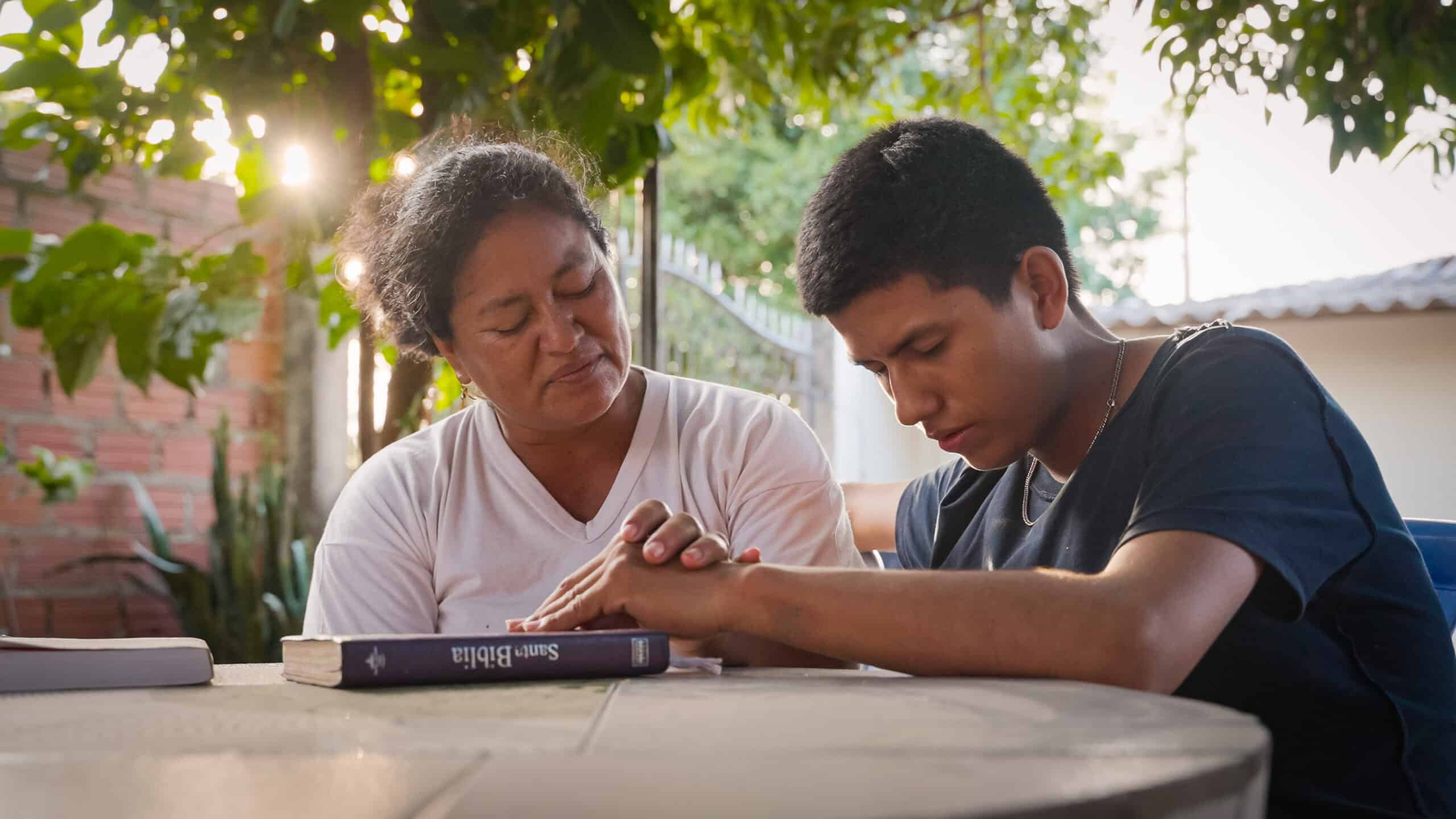 A young man sits with an older woman as they pray with their heads bowed and eyes closed.