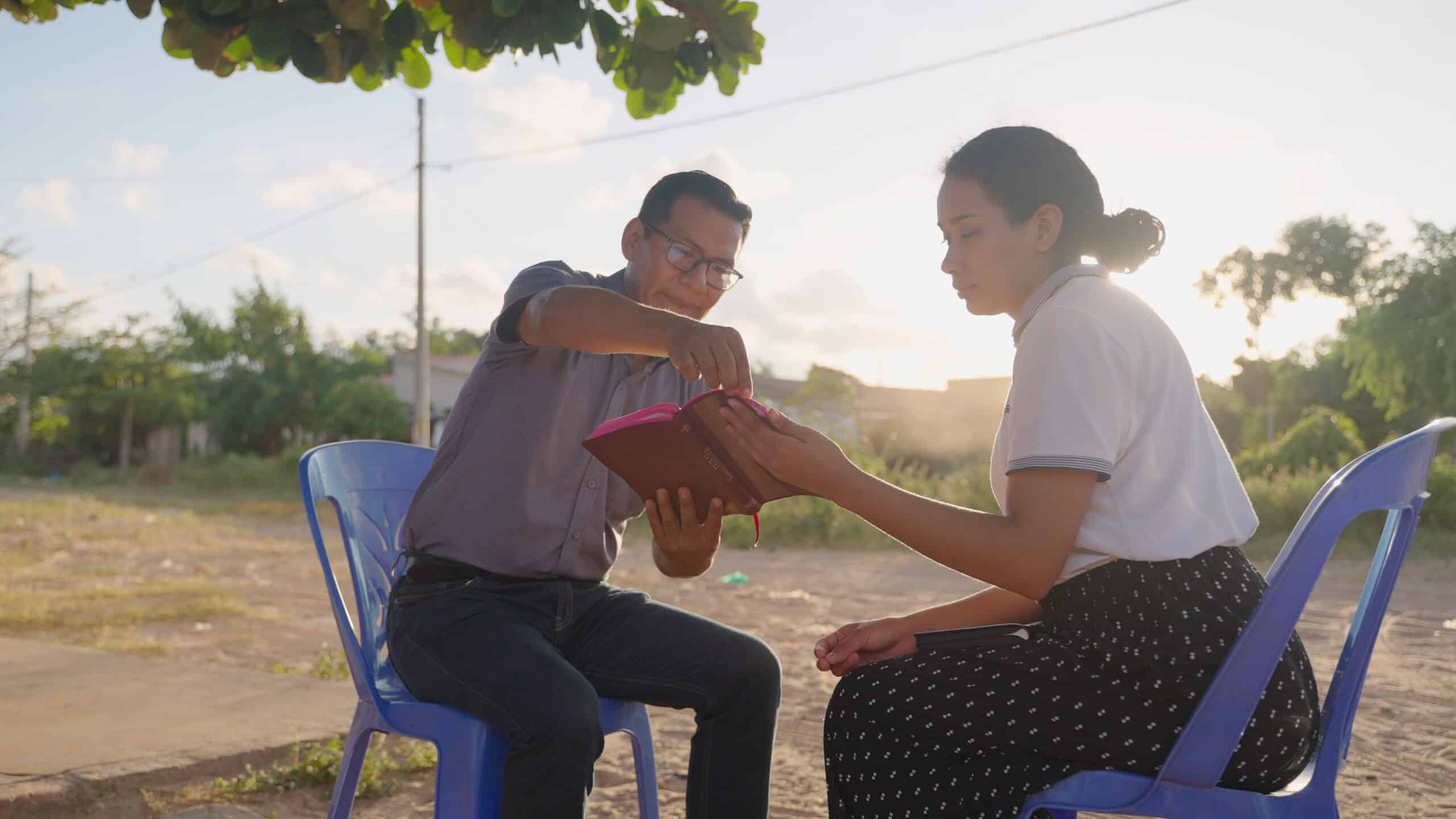 A young woman and an older man sit on blue chairs while holding a Bible between them and talking.