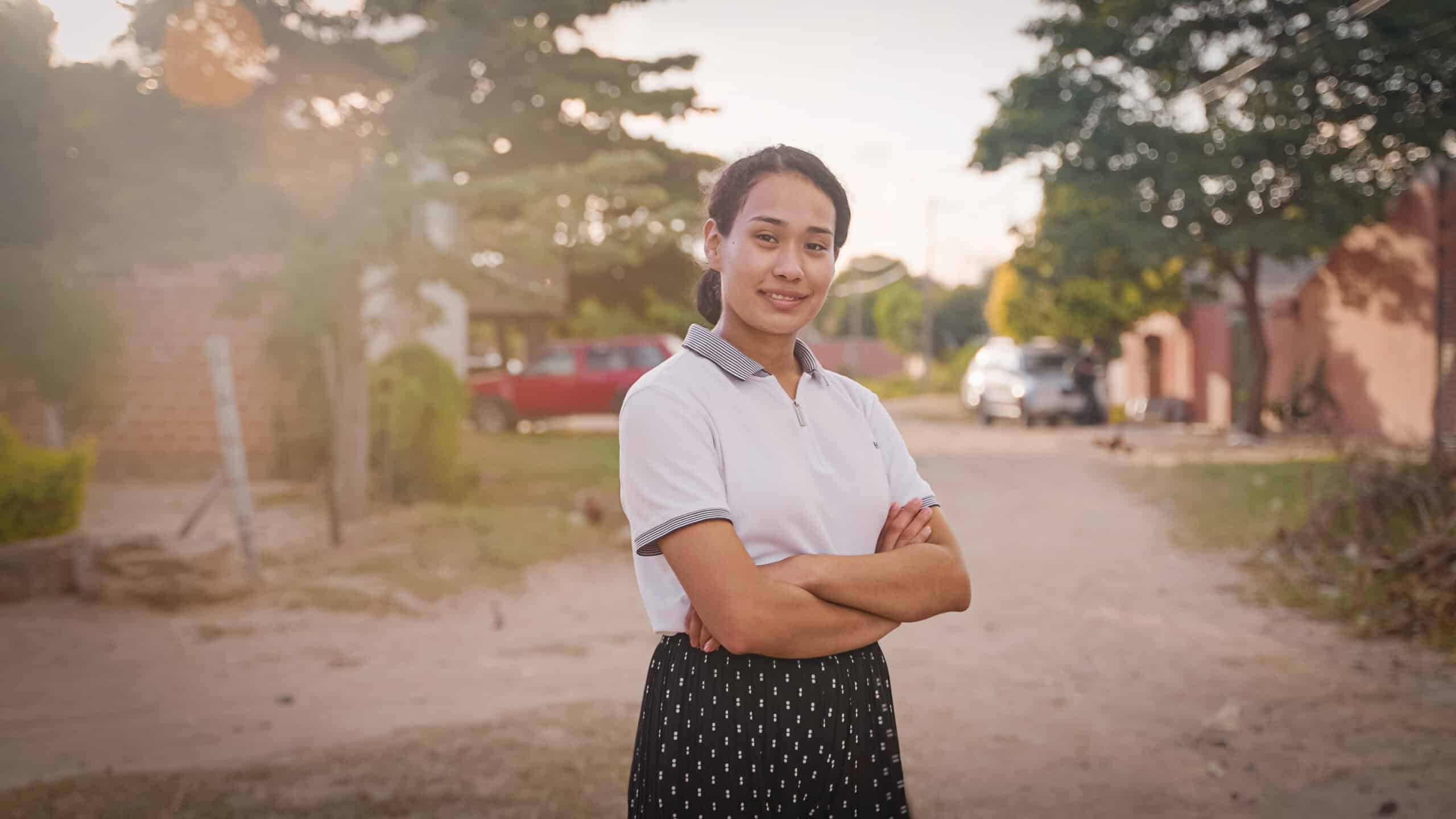 Young woman wearing a white collared shirt and block skirt stands with her arms crossed smiling. 