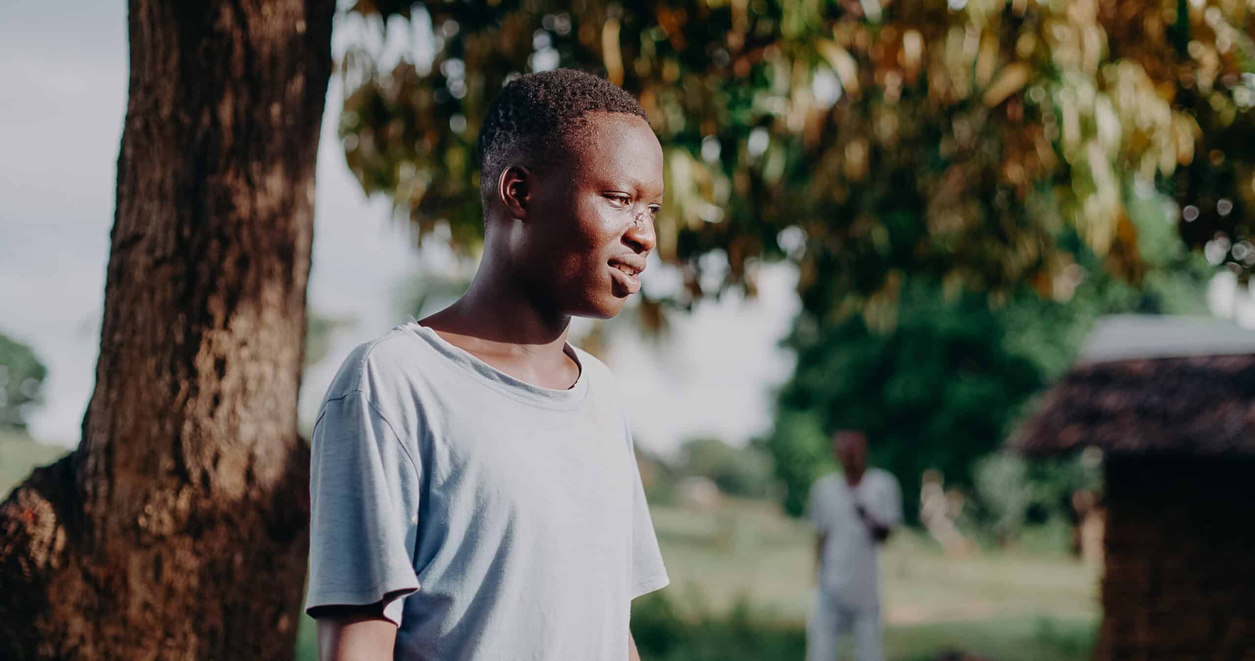 A young man in a blue t-shirt stands smiling and looking out into the distance.